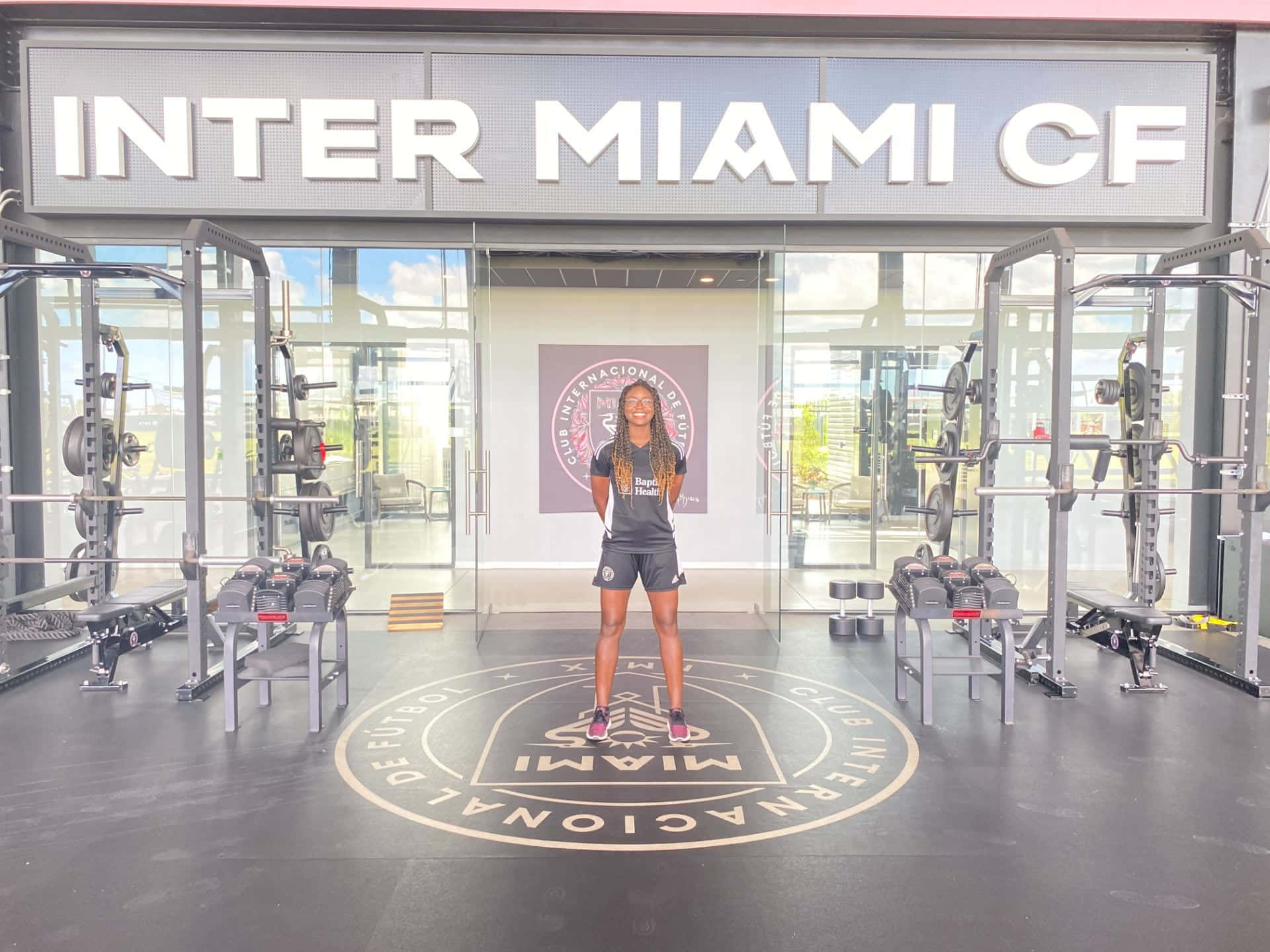 Inter Miami FC 訓練設施廣角鏡頭 壁紙 — 一位微笑的女性穿著運動服，自信地站在國際邁阿密CF的健身房中，舉重器材和球隊標誌清晰可見。
