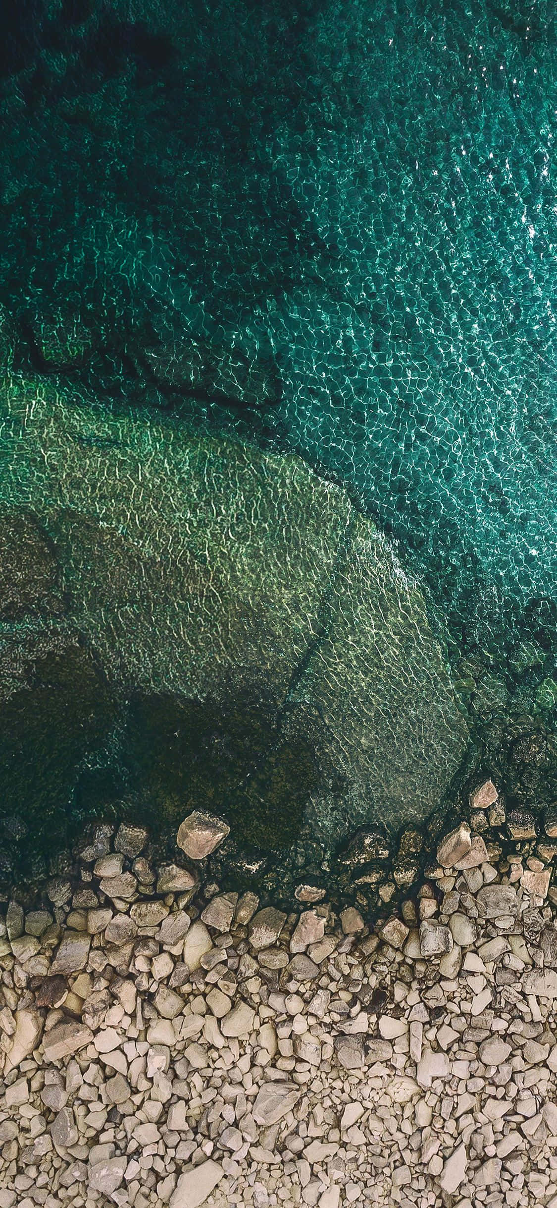 iOS 1 岩石海岸風景 壁紙 — 從高空俯瞰，清澈碧綠的海水在陽光照耀的岩石上泛起漣漪，形成岩石海岸線的景觀。