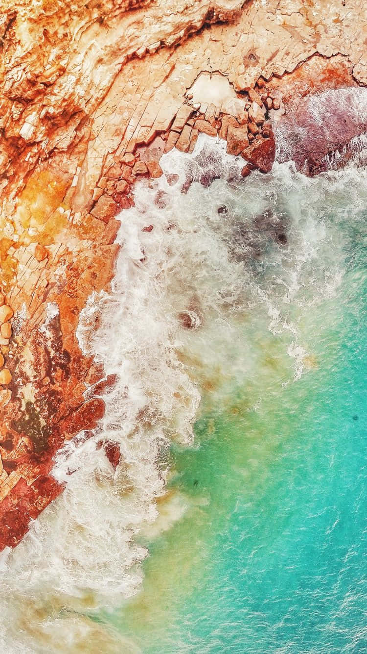iOS 1 浪花拍打棕色岩石 壁紙 — 從高空俯瞰，碧綠色的海浪拍打著崎嶇的橙色懸崖，構成戲劇性的海岸景觀。