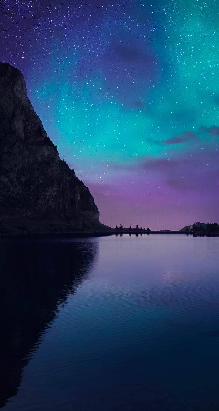 iOS 8 北極光 壁紙 — 繁星點點的夜空，充滿活力的綠色和紫色極光完美地映照在平靜的湖面上，旁邊是黑暗的岩石懸崖。