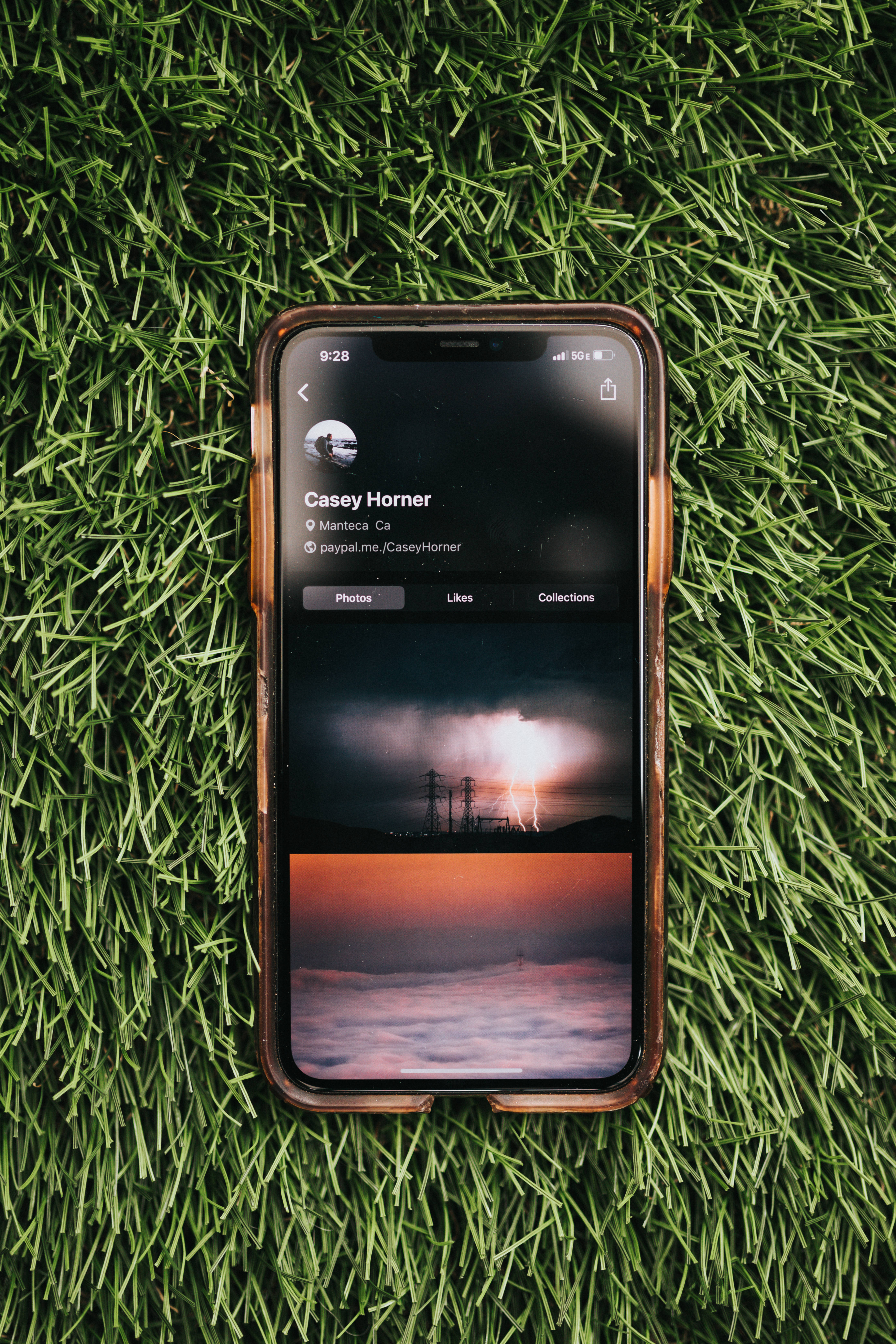 iOS iPhone 自然風景顯示 壁紙 — 一部帶有棕色保護殼的智能手機放在鮮綠色的草地上，顯示一個帶有暴風雨和日落照片的社交媒體個人資料。