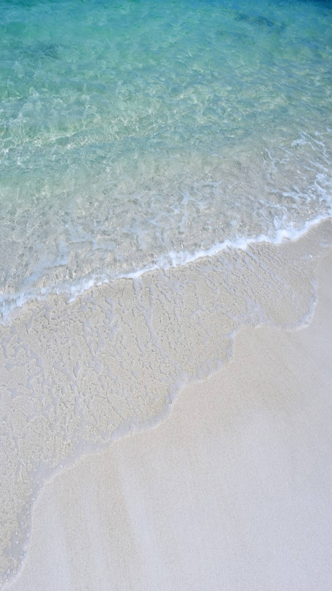 海灘岸邊 iOS預設 壁紙 — 清澈的碧綠海浪輕輕拍打柔軟的白沙，呈現寧靜的熱帶海灘景象。