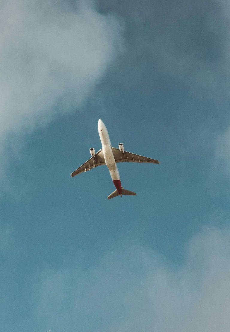 飛行客機 iPad 2021 壁紙 — 一架白色客機帶紅色尾翼在頭頂翱翔，背景是廣闊的藍天，點綴著柔軟的白雲。