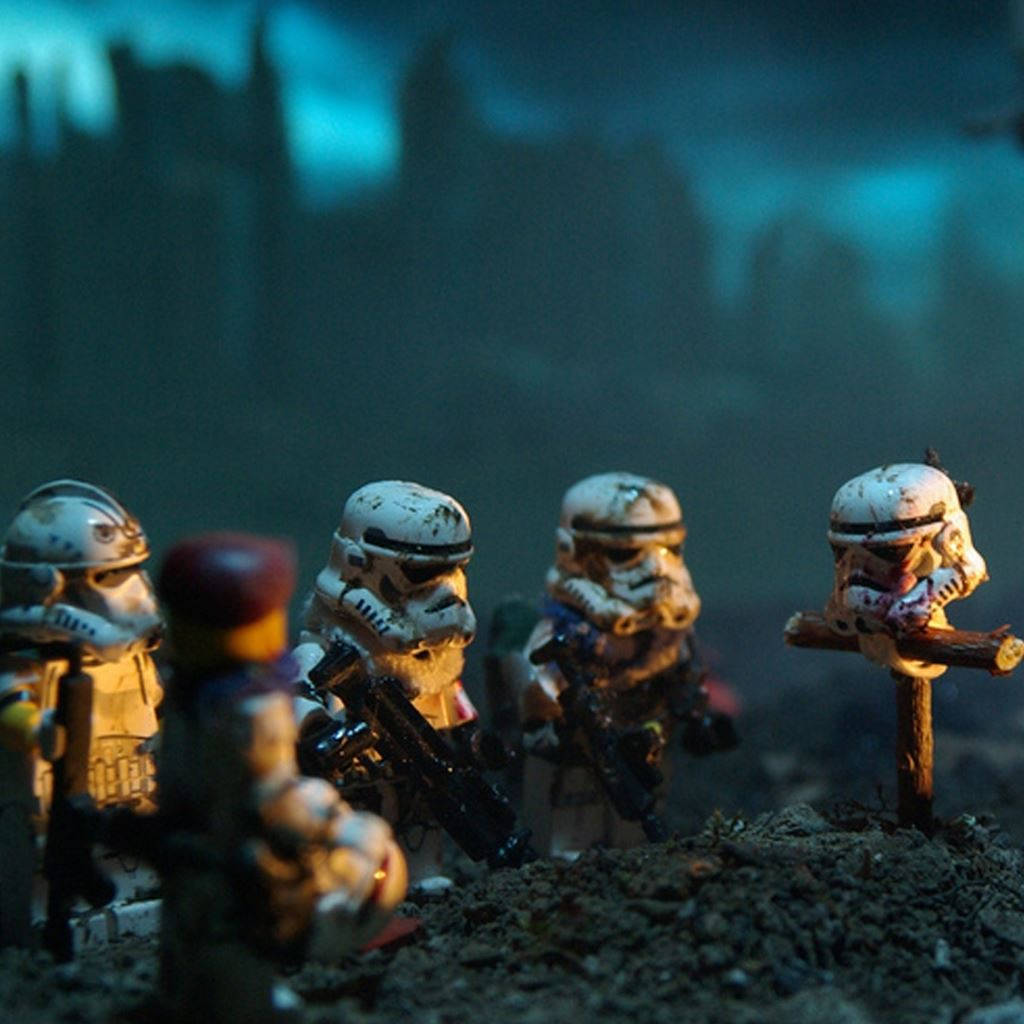 星際大戰 iPad 乐高風暴兵哀悼 壁紙 — 佈滿灰塵的LEGO風暴兵站在昏暗的戰場上，陰鬱的藍天下，一個倒下的同袍在臨時十字架上。