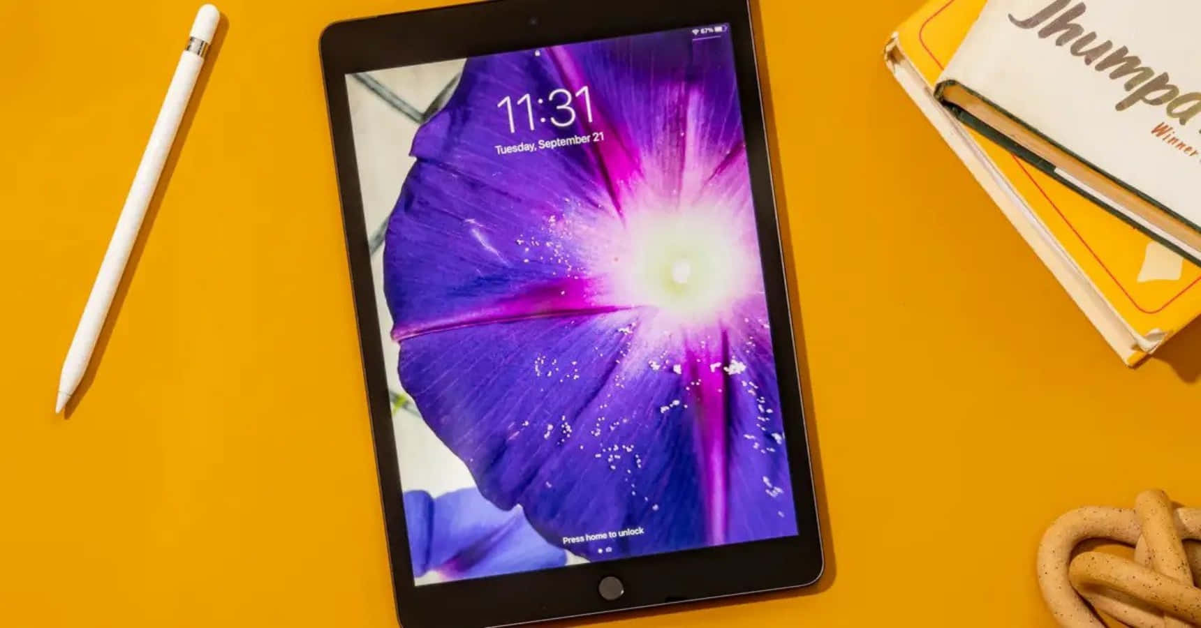 iPad Pro — 讓事事更高效 壁紙 — 一部顯示鮮豔紫色花朵桌面的平板電腦，放置在亮黃色表面上，旁邊有觸控筆和書籍。