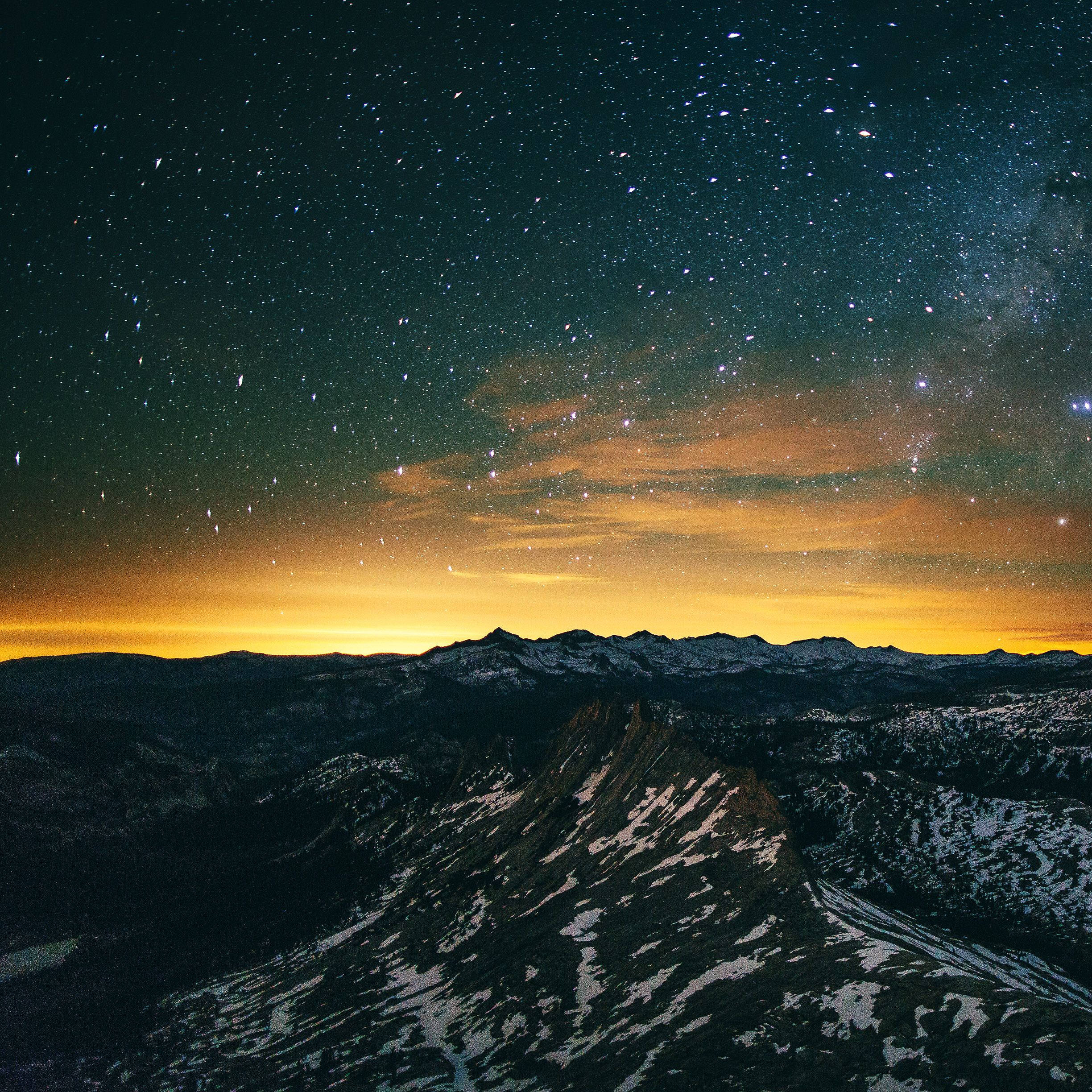 iPad Pro 山巒星夜夕照 壁紙 — 白雪覆蓋的山脈在繁星點點的夜空下升起，夜空閃爍著橙色極光雲。