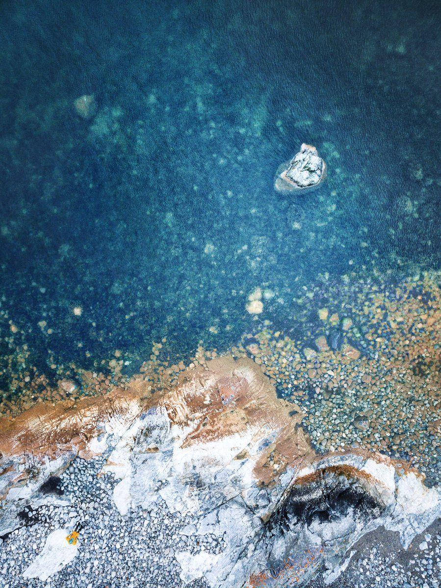iPad Pro 近小石灘的水域 壁紙 — 俯瞰視角，一塊孤獨的岩石漂浮在深藍色水域之上，下方是鏽色與白色鵝卵石組成的紋理海岸線。