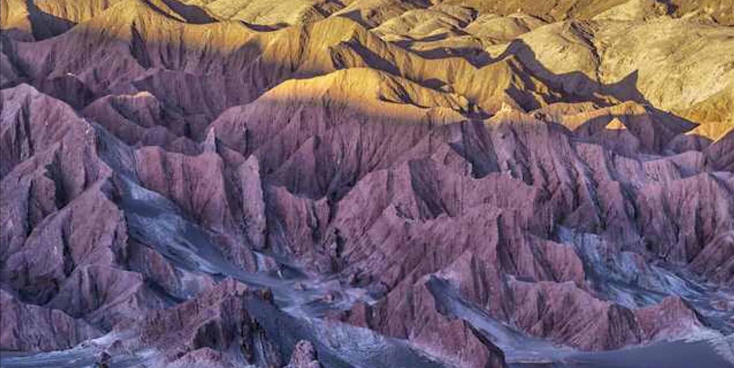 iPad Pro 阳光照耀的岩石山脈 壁紙 — 崎嶇峽谷的鳥瞰圖，紫色和金色的岩層沐浴在溫暖的日落光線中。