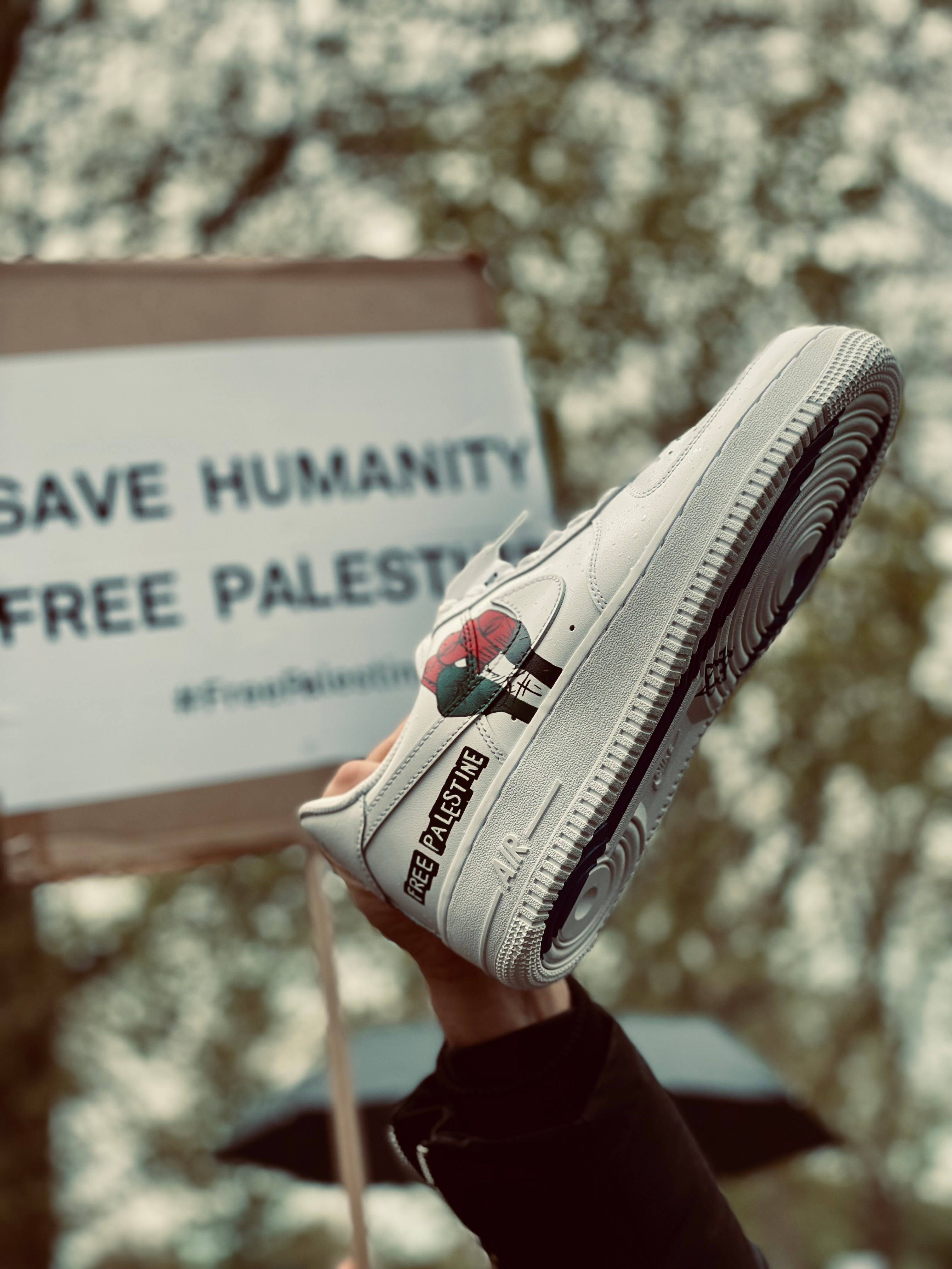 iPad Pro 鞋子舉起標語 壁紙 — 一隻手拿著一隻白色運動鞋，上面有 'FREE PALESTINE' 和一朵玫瑰補丁，位於抗議標語牌上方，標語牌寫著 'SAVE HUMANITY FREE PALESTINE'。