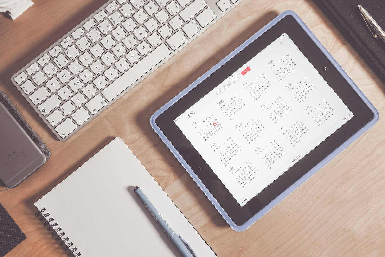 iPad Pro居家辦公場景 壁紙 — 木製書桌的俯視圖，桌上有一個顯示日曆的平板電腦、鍵盤、筆記本和筆，整齊排列以提高生產力。