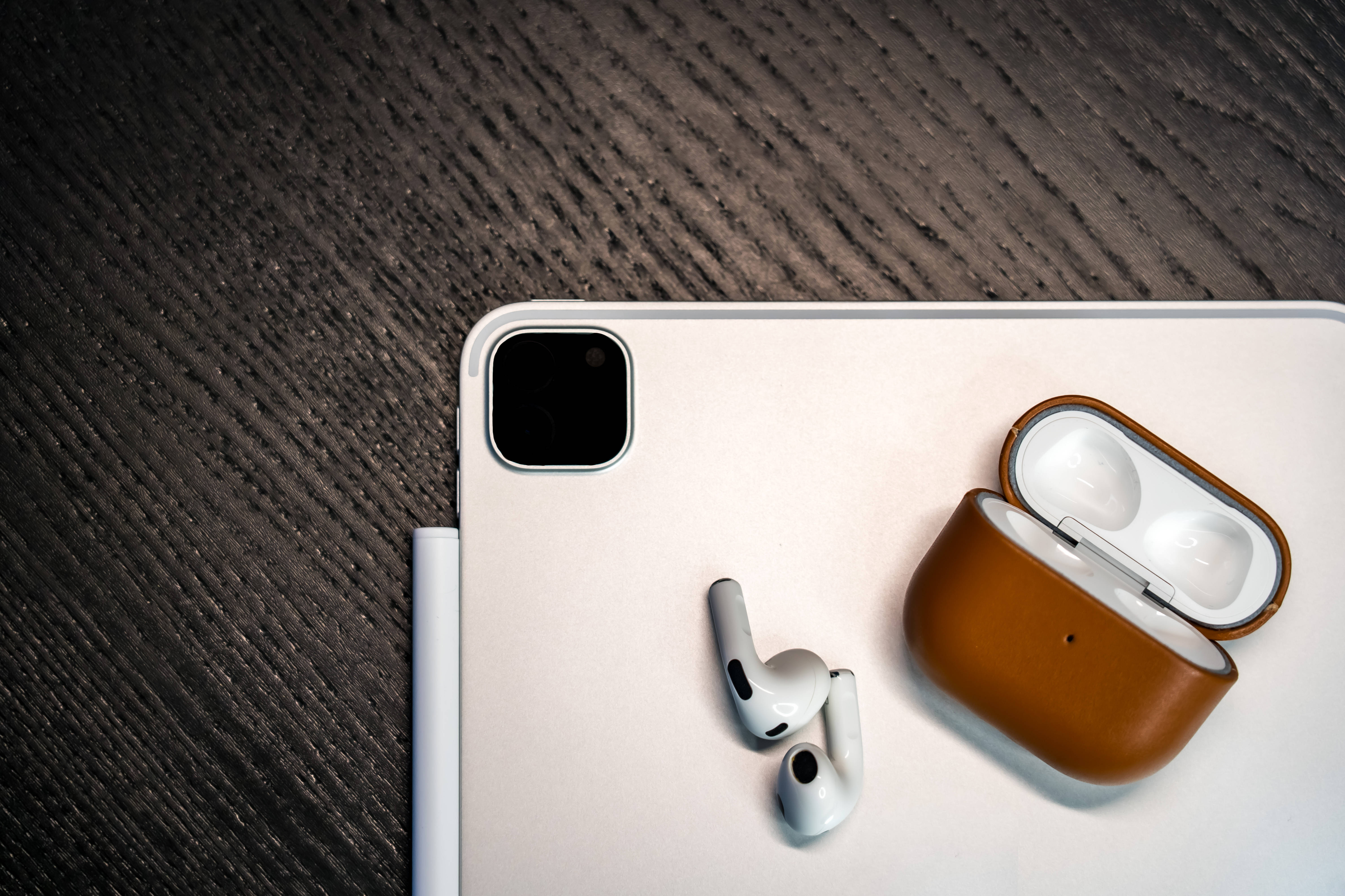 iPad Pro搭配AirPods 壁紙 — 白色無線耳機及其棕色皮革保護套放在深色木質表面上，旁邊有一部銀色平板電腦。