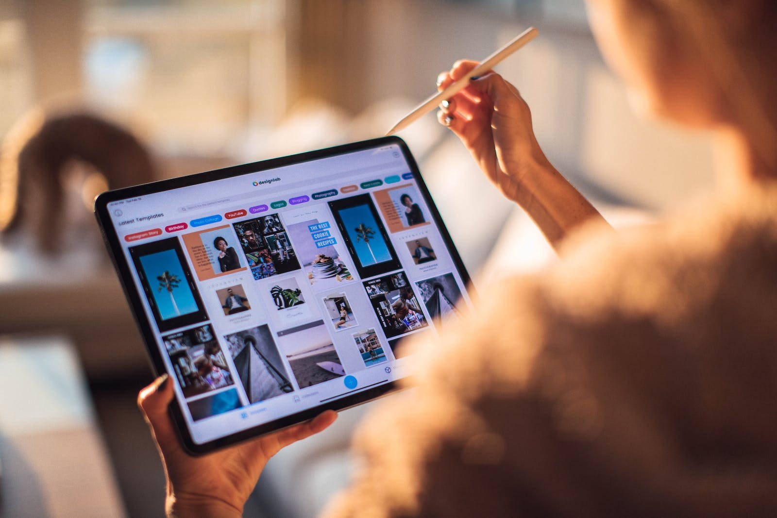 iPad Pro搭配Apple Pencil 壁紙 — 從肩膀上方視角，看到一個人在光線溫暖的室內空間中使用平板電腦瀏覽設計模板。