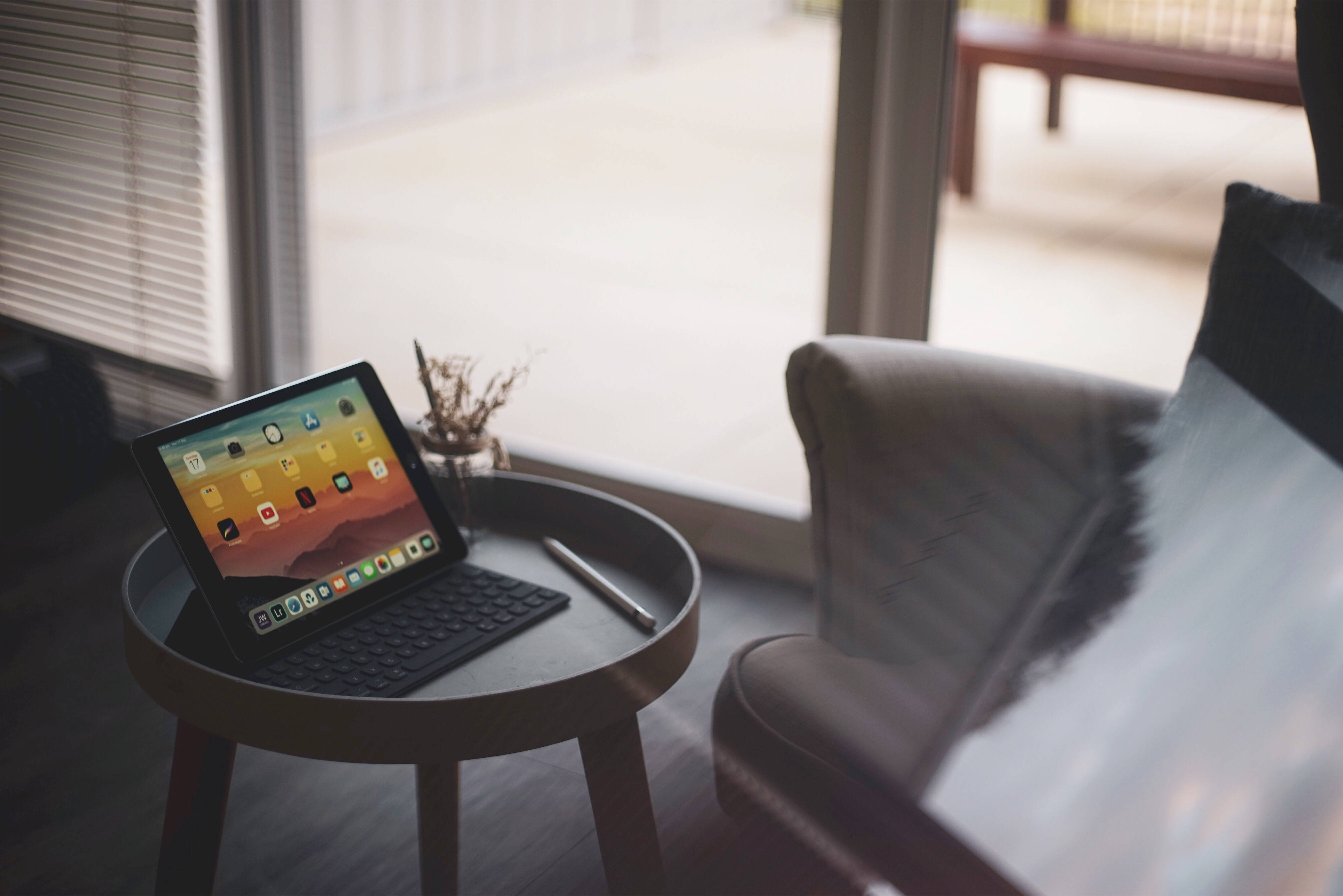 iPad Pro置於咖啡桌 壁紙 — 一部帶鍵盤的平板電腦放在木製邊桌上，旁邊是一張舒適的扶手椅，靠近帶百葉窗的玻璃門。