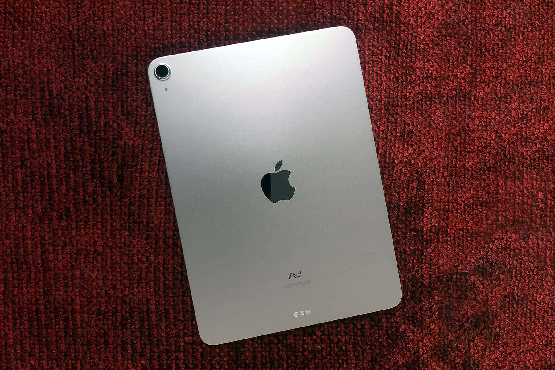 在最新iPad上享受您最愛的內容 壁紙 — 銀色iPad對角放置在質感豐富的紅色地毯上，帶有柔和環境光線。