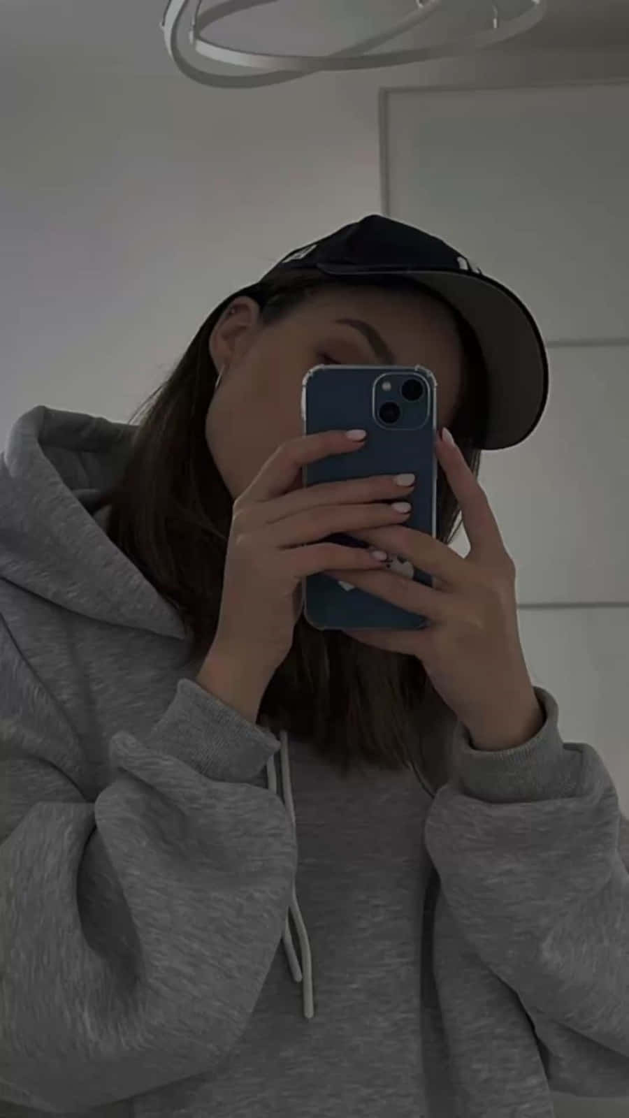 具抽象圖案的美學iPhone 壁紙 — 一個穿著灰色連帽衫和黑色帽子的人，在昏暗的室內燈光下用藍色手機拍鏡子自拍。