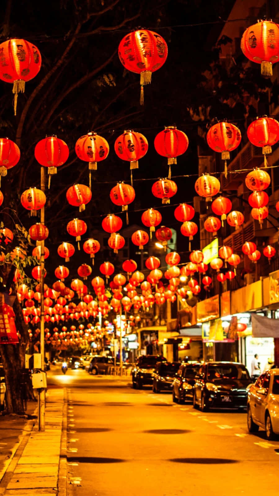 以最新iPhone慶祝中國新年 壁紙 — 一條充滿活力的夜街，掛滿了發光的紅燈籠，在停泊的汽車和樹木成蔭的人行道上投下溫暖的光芒。