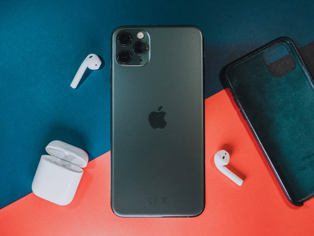 iPhone 11 Pro 磨砂黑 壁紙 — 一部時尚的黑色 iPhone，配以白色 AirPods 和保護殼，排列在鮮明的青綠色和珊瑚色背景上。
