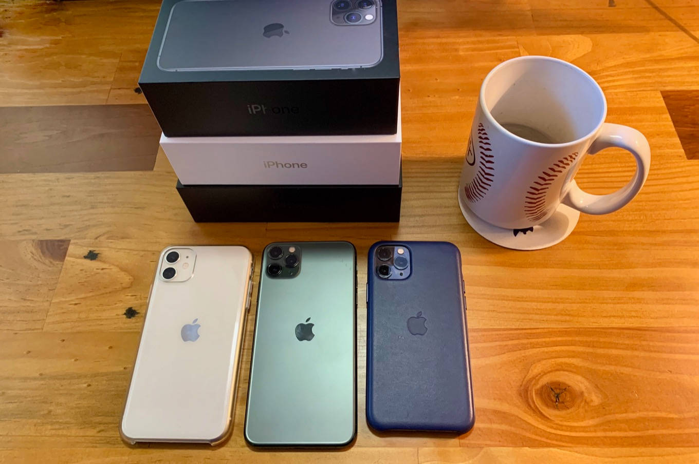 iPhone 11 放置於木製桌面 壁紙 — 三部白色、綠色和藍色的iPhone放在木桌上，旁邊有它們的盒子和一個棒球杯。