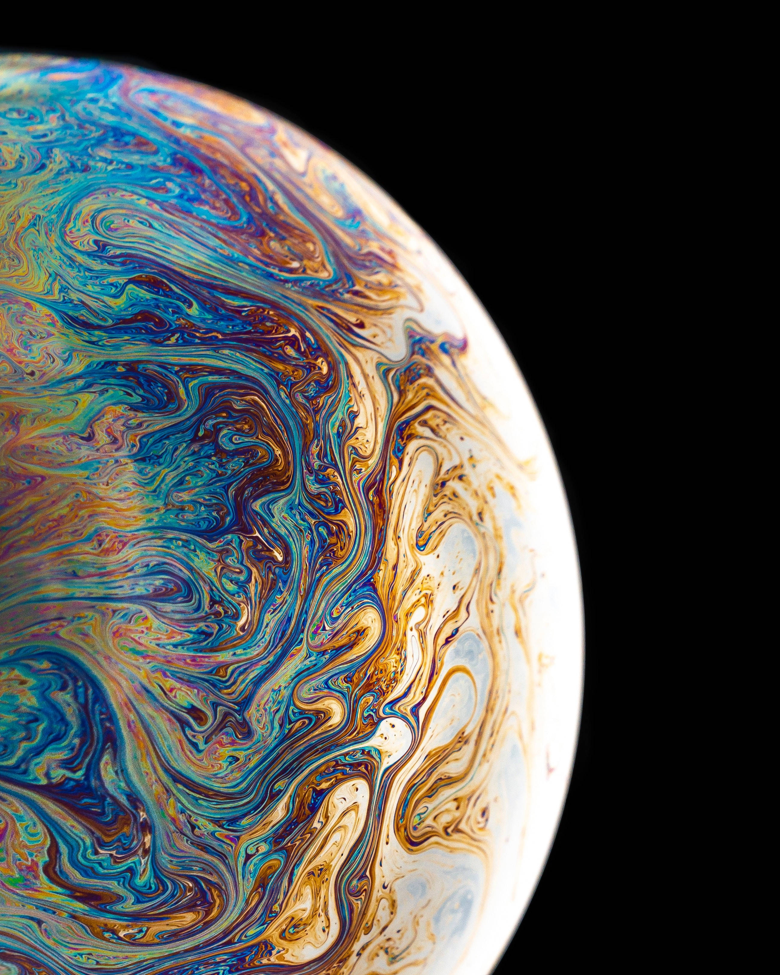 iPhone 12 Pro Max 迷幻星球 壁紙 — 一個肥皂泡的虹彩表面特寫，在純黑背景下旋轉著鮮豔的藍色、金色和白色。