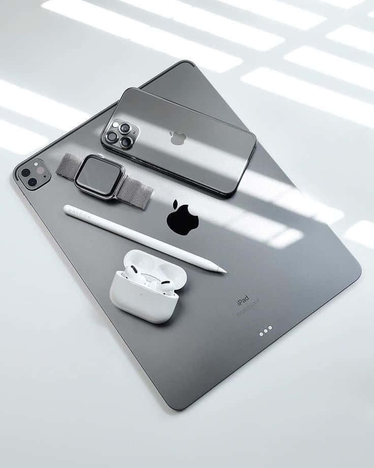iPhone 12 將 5G 速度握在掌心 壁紙 — 現代Apple裝置包括iPad、iPhone、Apple Watch和AirPods，排列在時尚的灰色表面上，明亮的窗戶光線投射出幾何陰影。