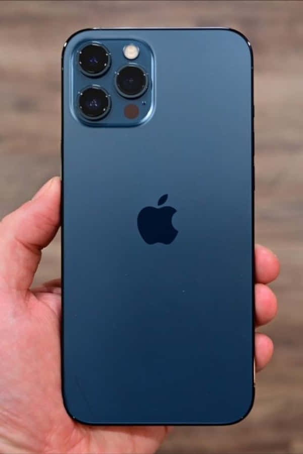 iPhone 12：感受 5G 的差異 壁紙 — 一隻手拿著一部時尚的藍色iPhone，帶有突出的相機模組和Apple標誌，背景是模糊的木質背景。