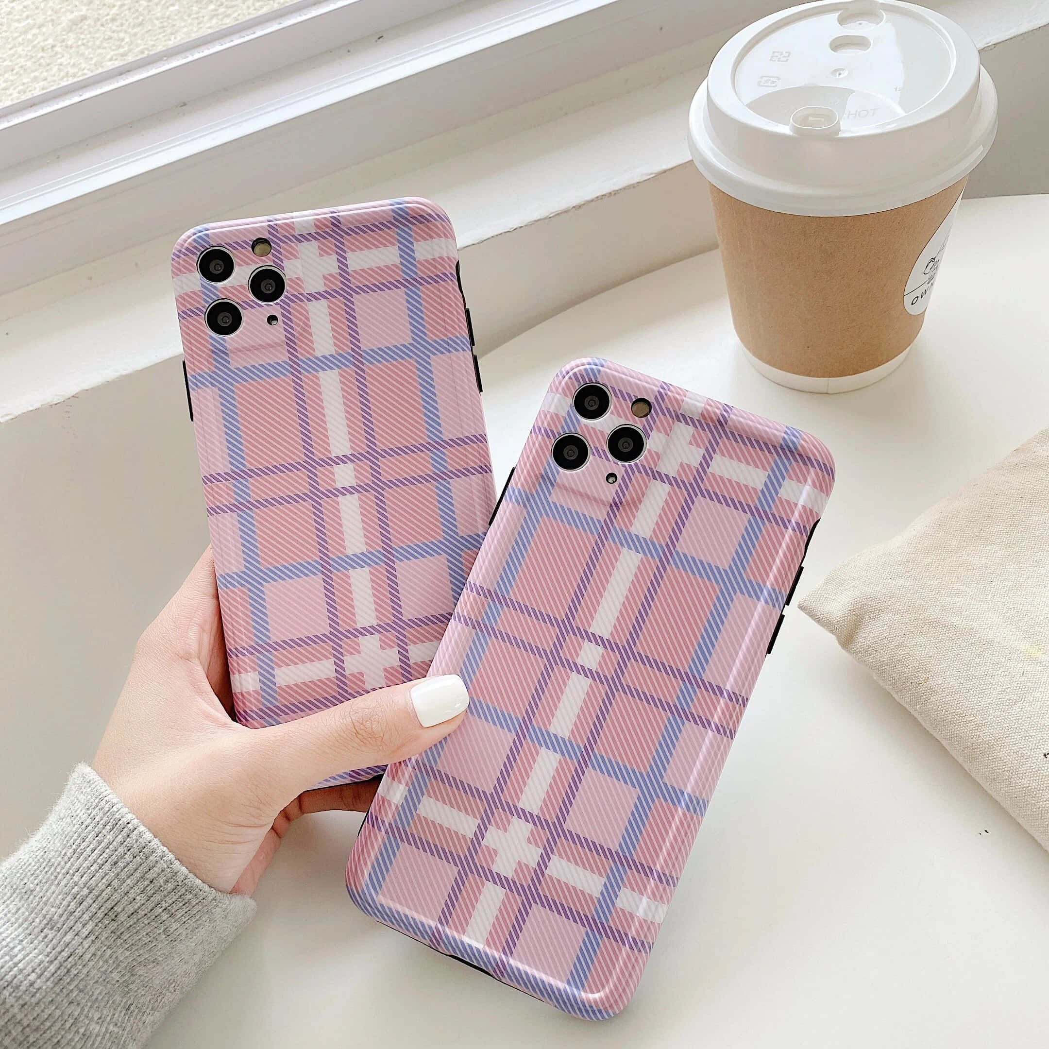 可愛時尚的iPhone 壁紙 — 一隻手拿著兩個粉紅、紫和白格仔圖案的手機殼，放在窗台上的咖啡杯旁邊。
