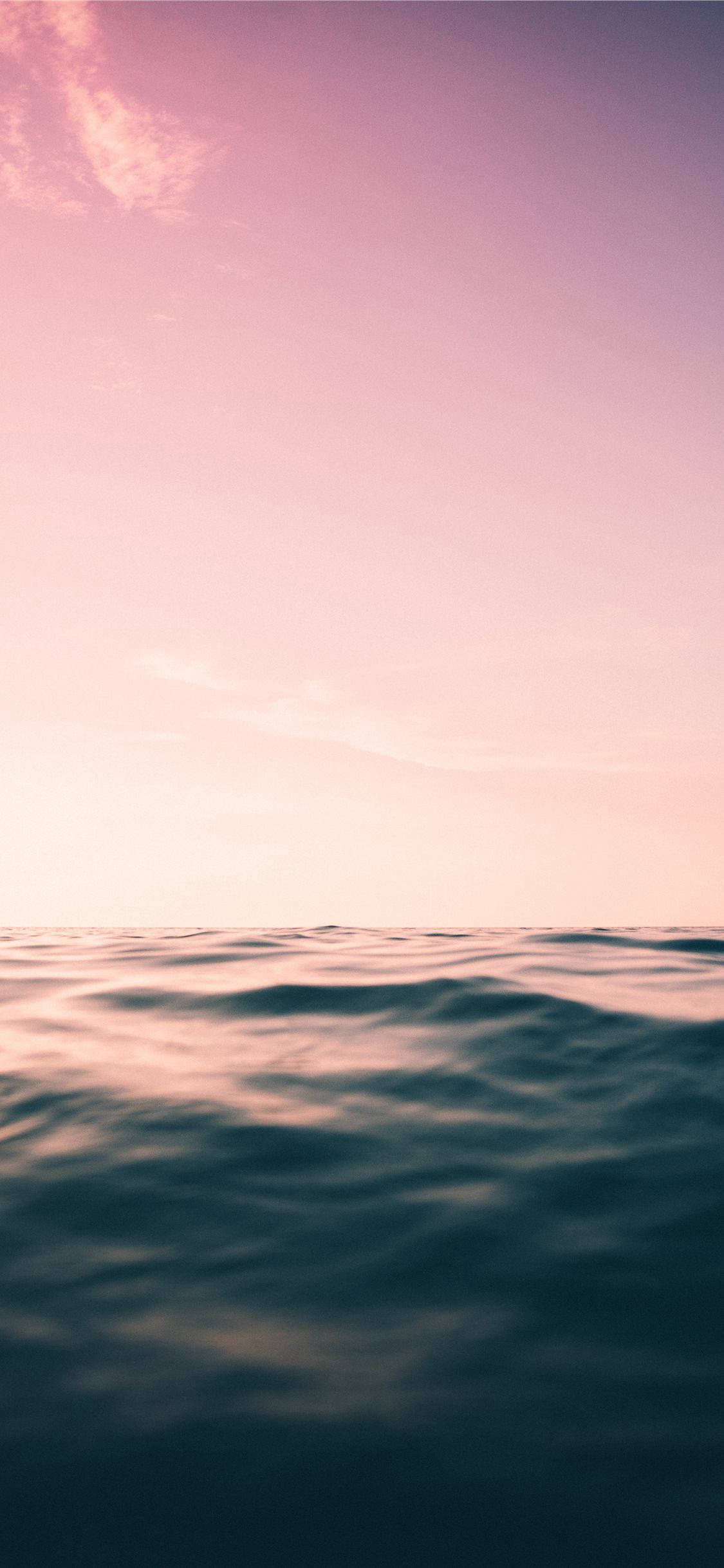 iPhone 用的美學海洋水景壁紙 壁紙 — 寧靜的海平線沐浴在柔和的粉紅色和紫色暮色中，下方有輕柔的波浪。