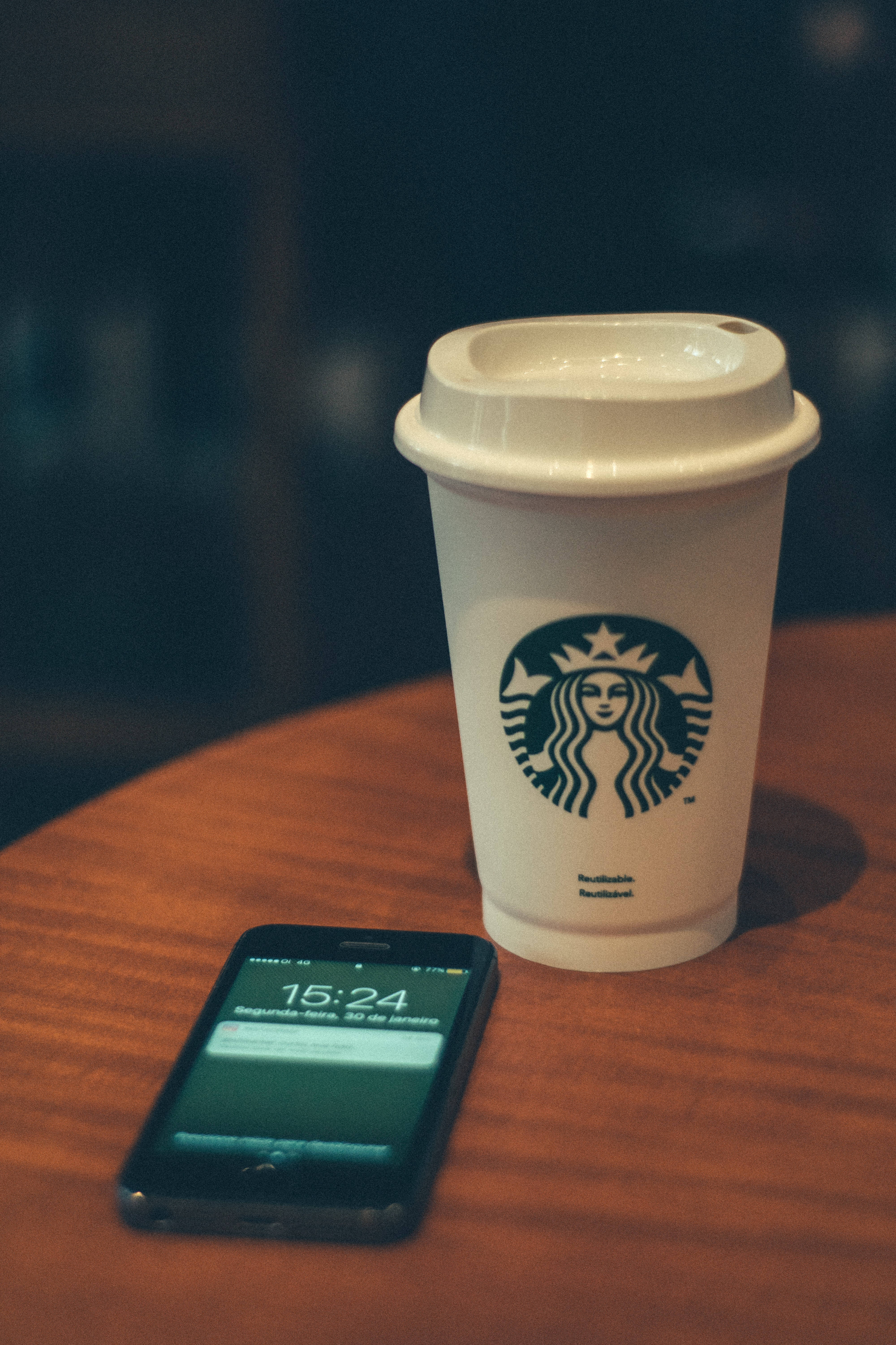 iPhone 4K 屏幕上的迷人城市夜景 壁紙 — 一個白色Starbucks杯和黑色智能手機放在溫暖的木桌上，背景是柔和模糊的咖啡館環境。