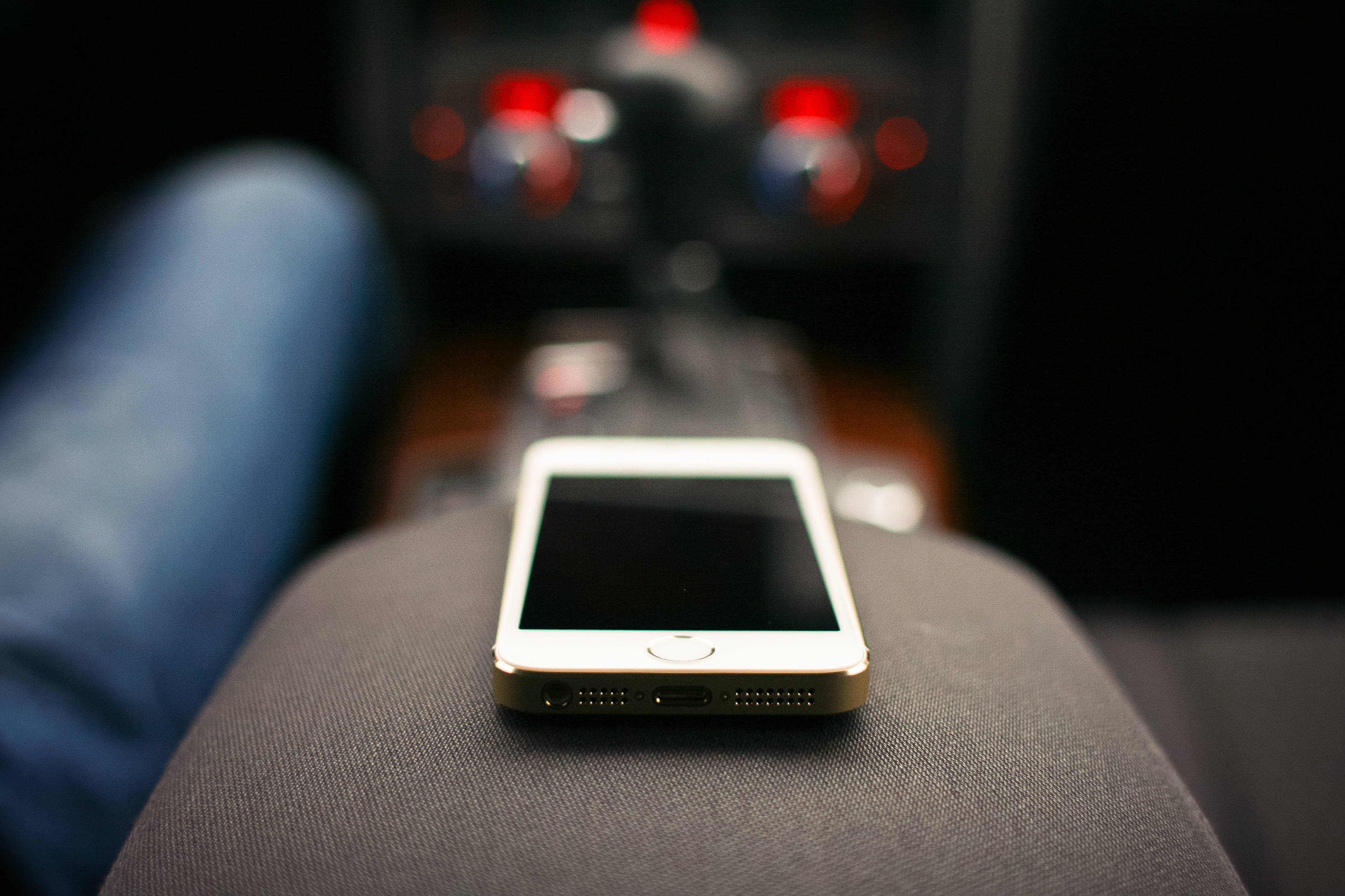 時尚優雅的原裝 iPhone 5 壁紙 — 一部金色智能手機放在一個人的大腿上，背景有模糊的汽車控制裝置和紅燈。