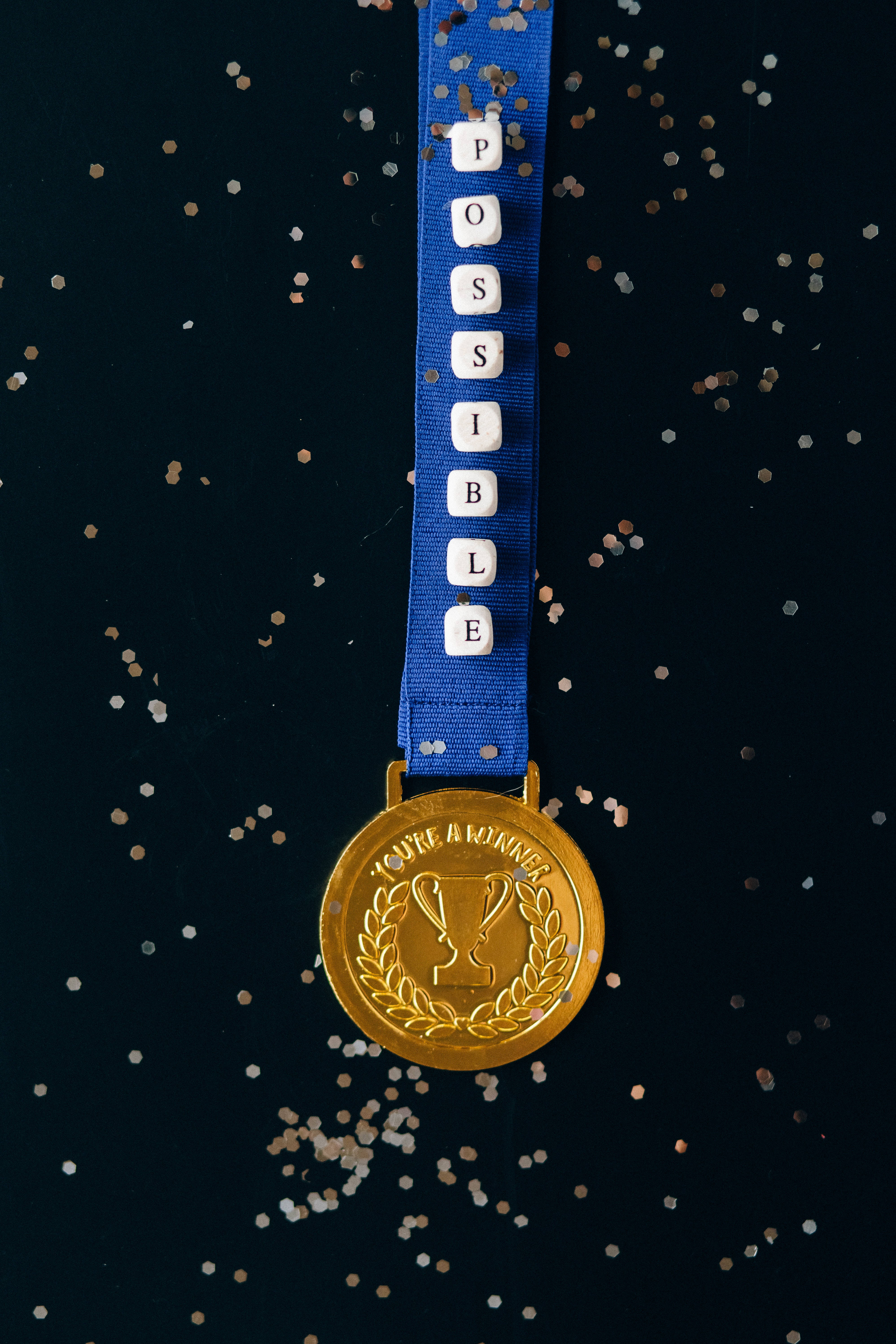 可能獎牌黑金iPhone壁紙 壁紙 — 一面金色獎牌，上面有「YOU'RE A WINNER」和月桂花環，掛在一條藍色絲帶上，拼出「POSSIBLE」，背景為深色並帶有閃粉。
