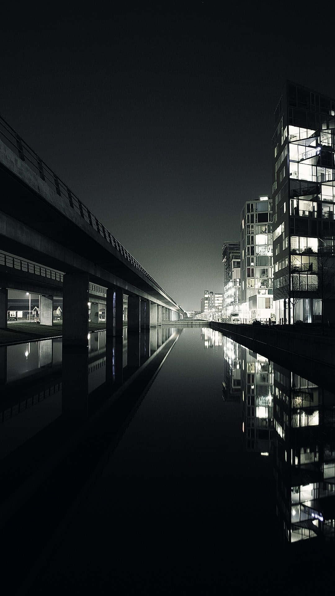 現代iPhone芝加哥夜景湖畔 壁紙 — 一條運河的單色夜景，兩旁是現代玻璃建築和高架橋，發光的窗戶倒映在平靜的水面上。