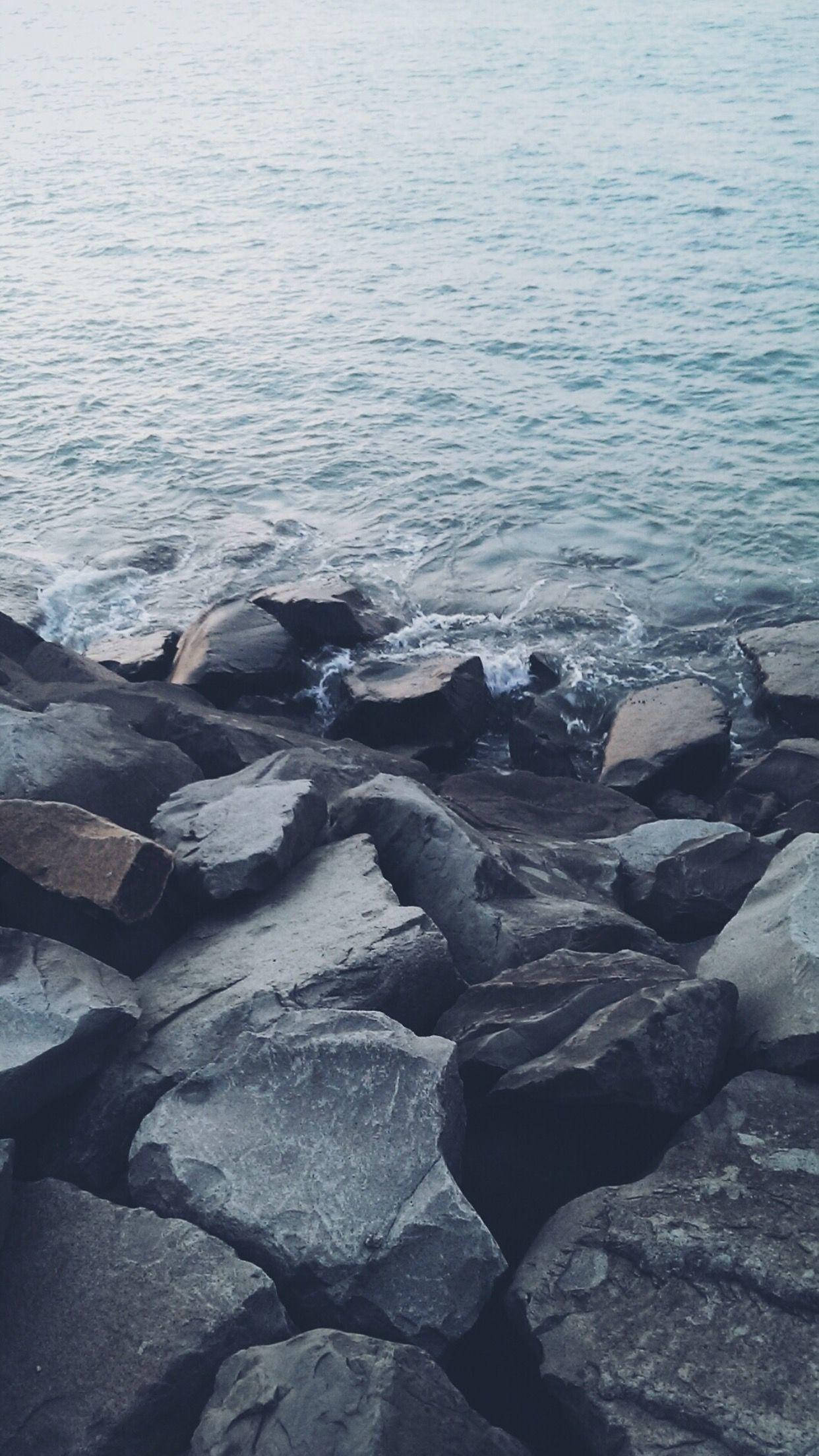 黑色與灰色海石 iPhone 6s Plus 壁紙 — 在陰沉天空下，海浪拍打著海岸線的深灰色岩石。
