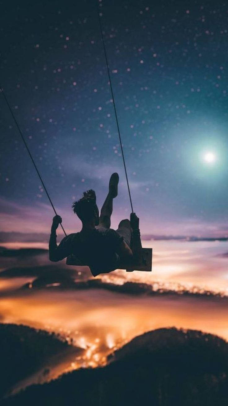 一個人在星光燦爛的夜空下，在發光的山谷上方盪鞦韆的剪影。