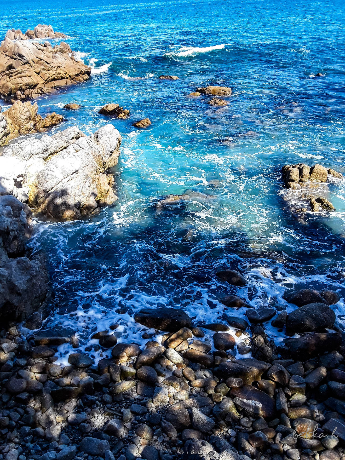 碧綠的海浪拍打岩石海岸，陽光下的鵝卵石閃閃發光。