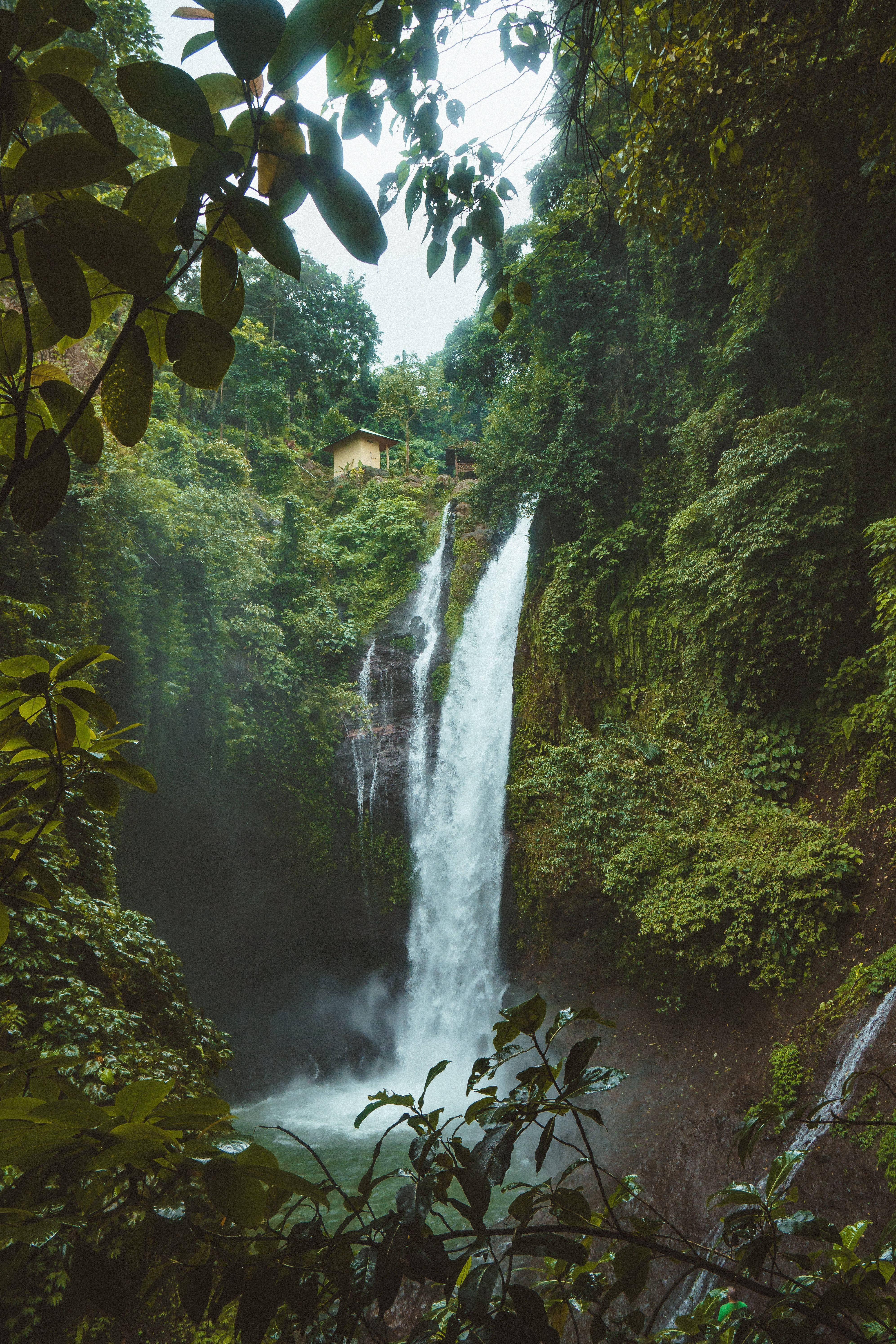 涼爽山景 iPhone 解鎖畫面 壁紙 — 一道雄偉的瀑布穿過茂密的綠色叢林，薄霧升起，懸崖邊上有一座小木屋。