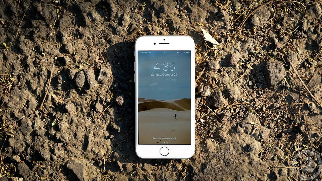 iPhone 8 陽光下 壁紙 — 一部白色智能手機放在粗糙的泥土地上，顯示沙漠壁紙，其中一個孤獨的人影在沙丘之間行走。