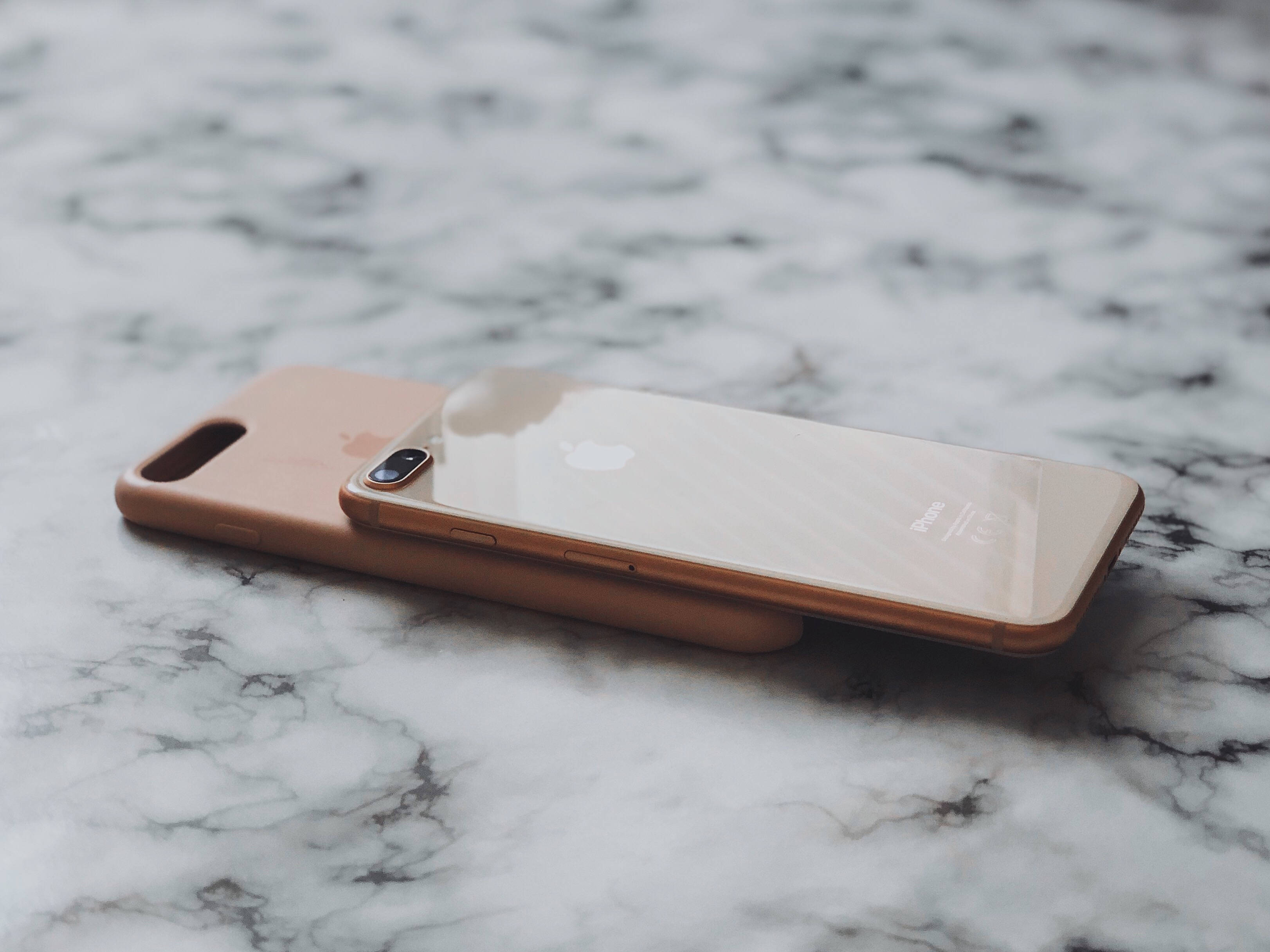 iPhone 8 加保護殼 壁紙 — 玫瑰金色iPhone放在相配的保護殼上，置於白色和灰色大理石表面，柔和自然光線照射。