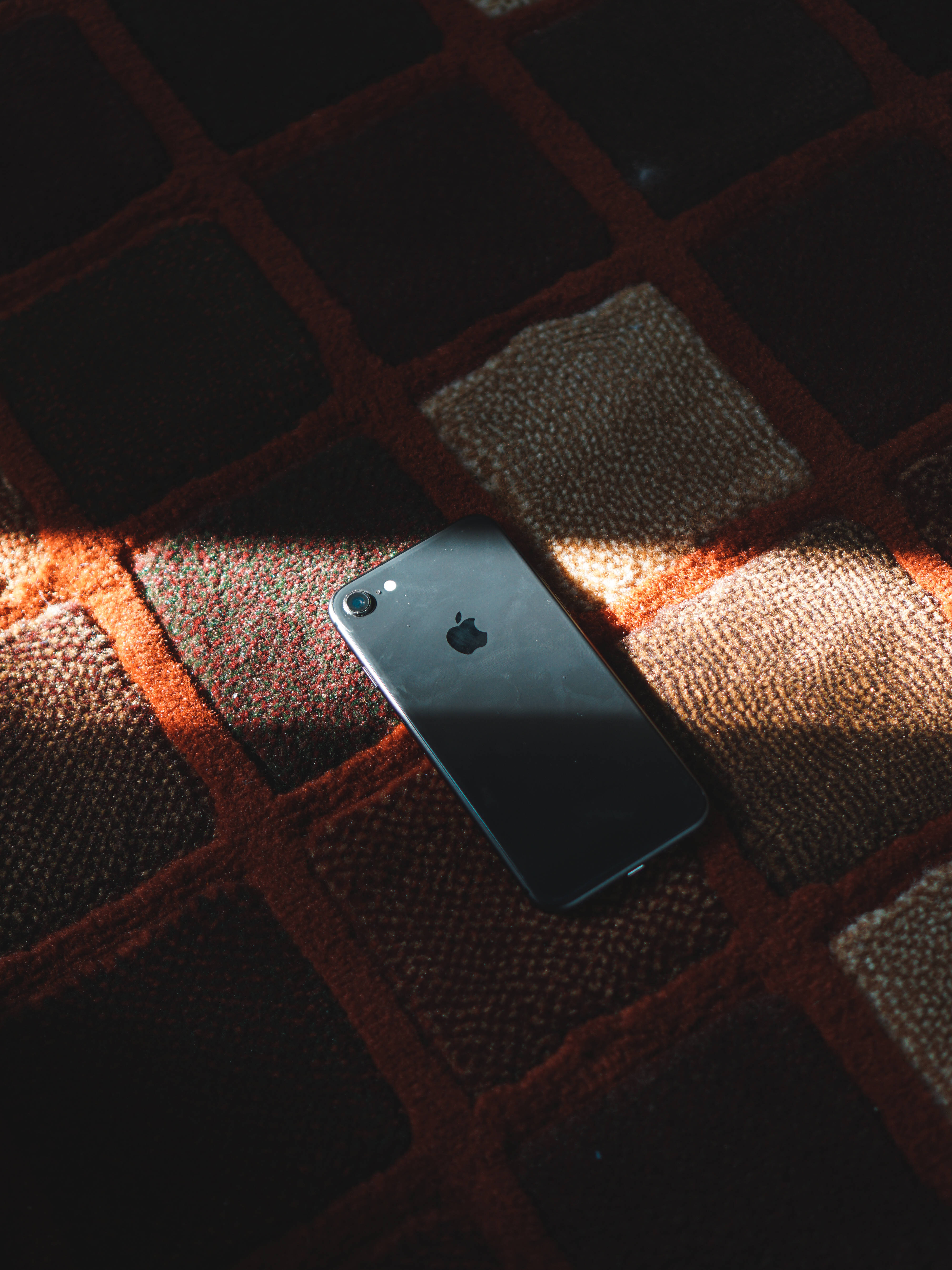 iPhone 8 陰影 壁紙 — 一部時尚的黑色iPhone放在一張質感豐富的紅米色格紋地毯上，沐浴在溫暖的陽光中。