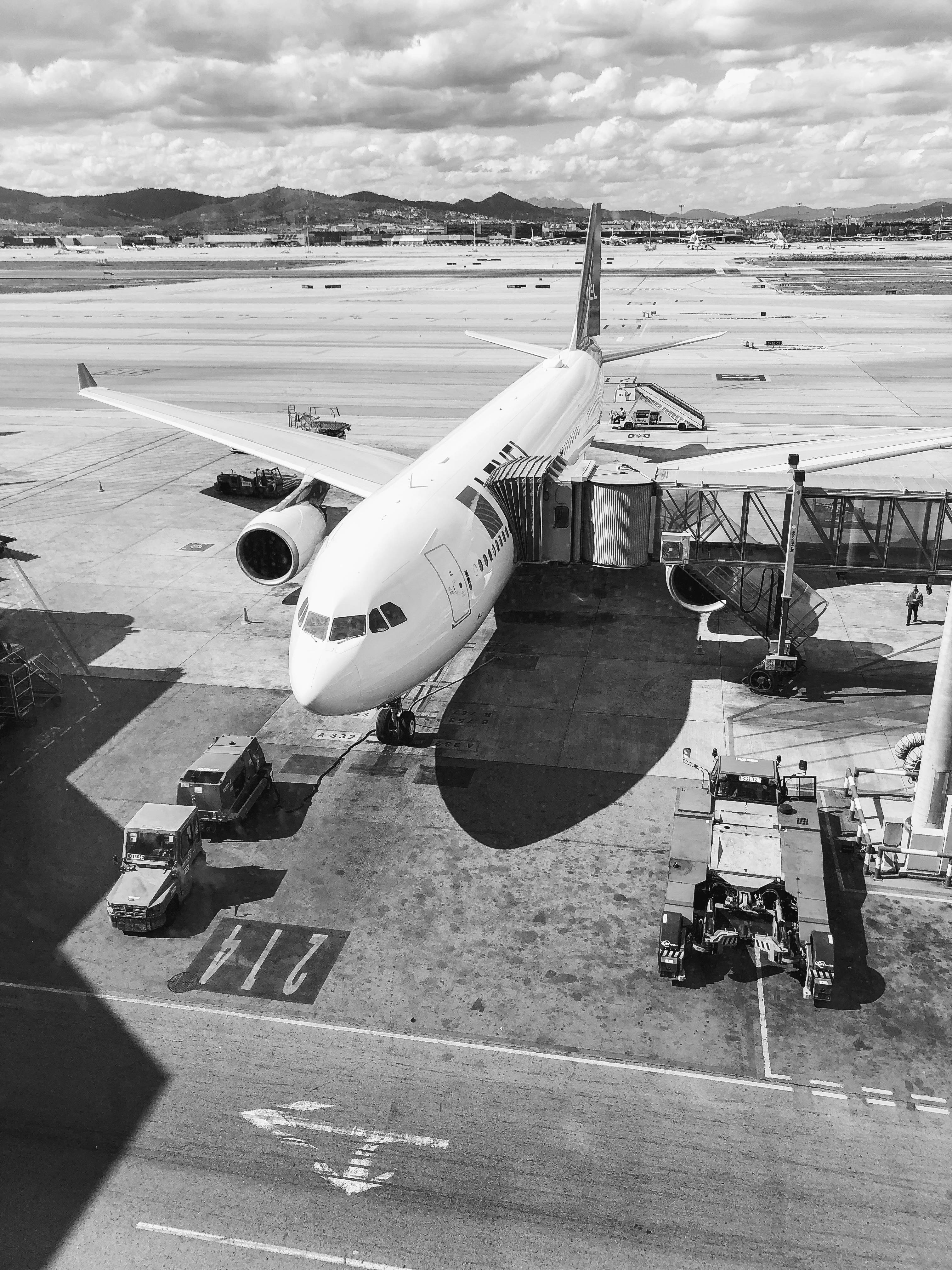 黑白飛機 iPhone 壁紙 — 黑白高角度視角，一架大型客機停泊在機場閘口，地面服務車輛和遠山在陰天背景下。