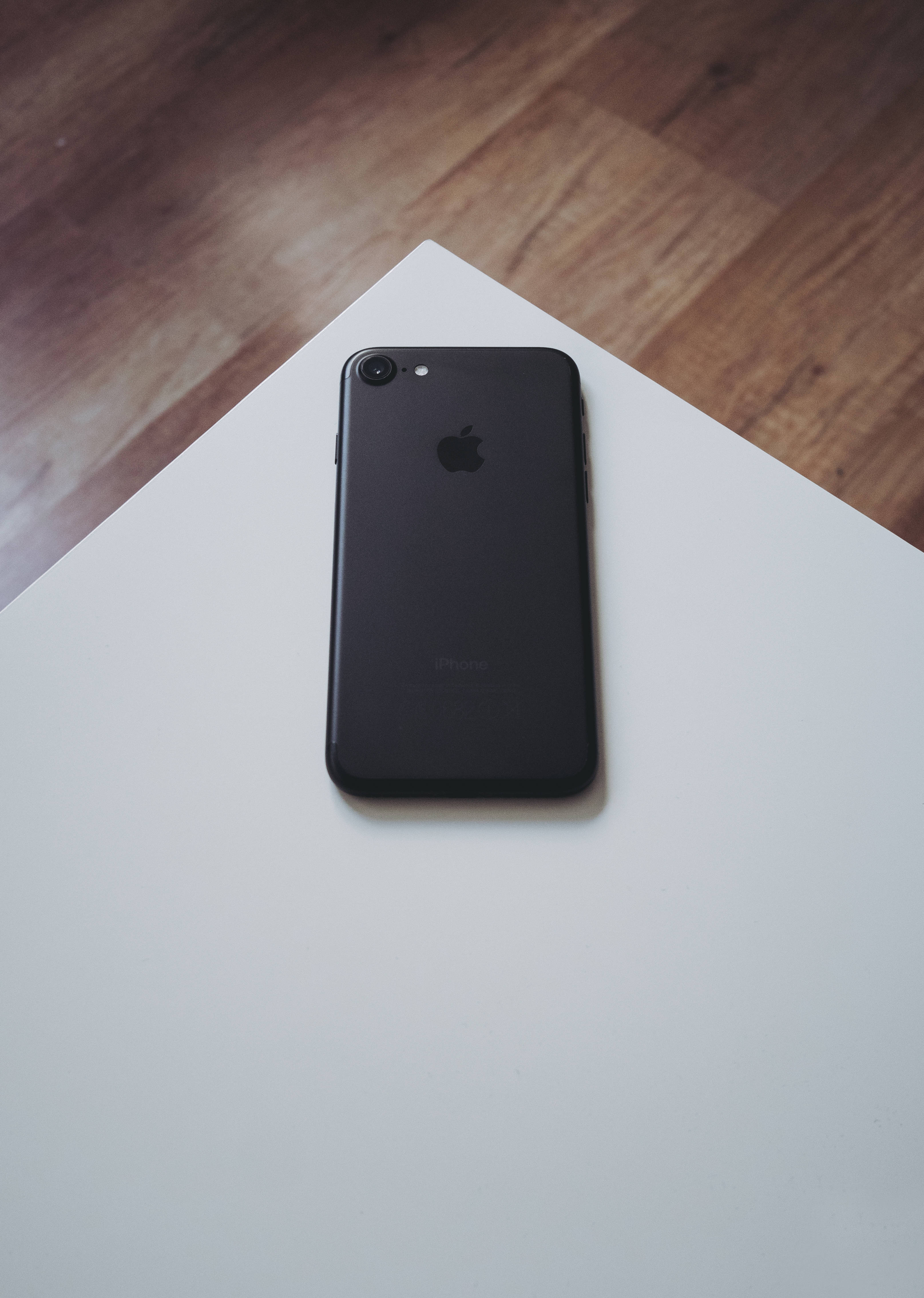 iPhone 8白色表面 壁紙 — 一部黑色iPhone 7放在白色表面上，背景是溫暖的木地板。