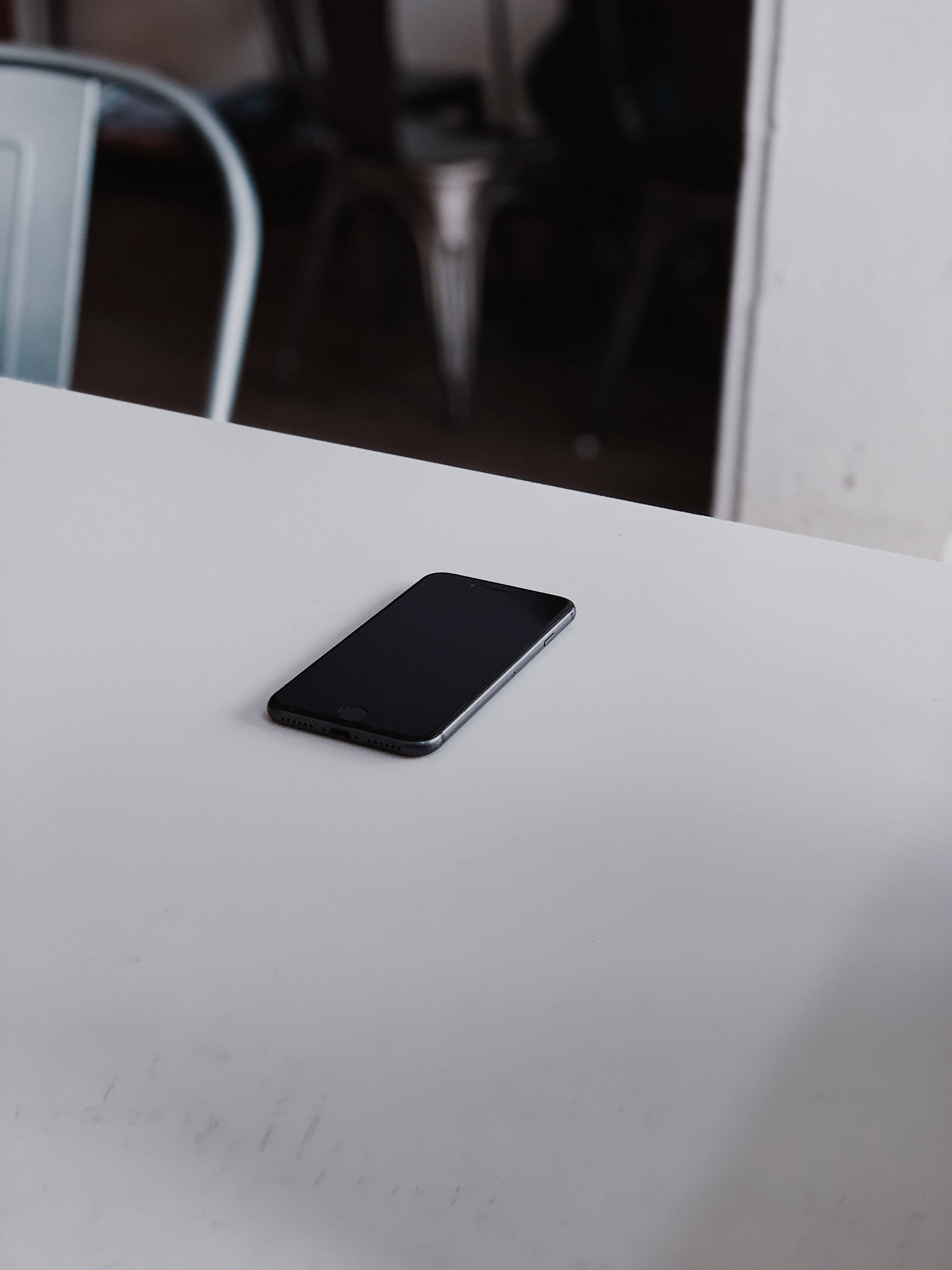 iPhone 8置於桌面 壁紙 — 一部時尚的黑色智能手機獨自放在簡約的白色桌子上，背景有一張模糊的金屬椅子。