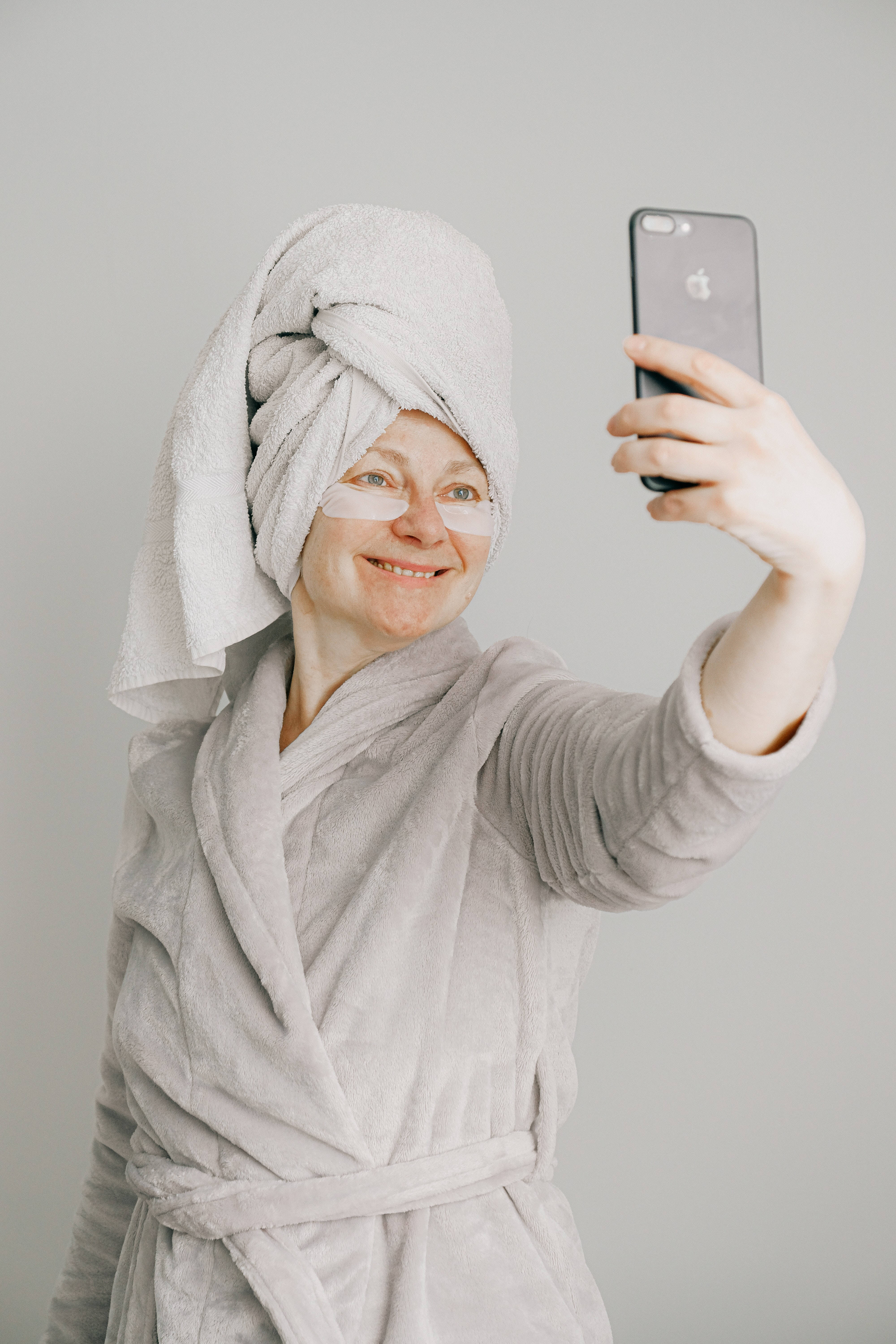 淋浴後的iPhone迷人自拍 壁紙 — 一位微笑的女子穿著灰色長袍，頭上裹著毛巾，貼著眼膜，在純灰色背景前用手機自拍。