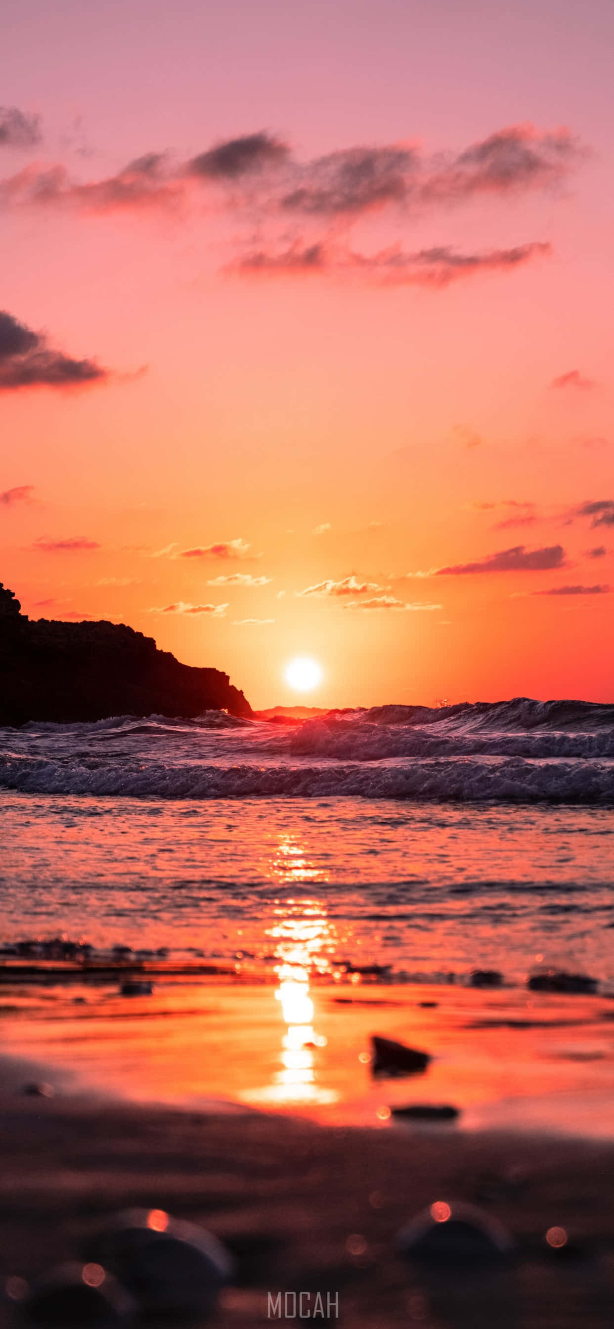 陽光灑落的海灘日出，完美 iPhone 風景壁紙 壁紙 — 充滿活力的夕陽將天空和海洋染成火熱的橙色和粉紅色，海浪拍打著岩石海岸。