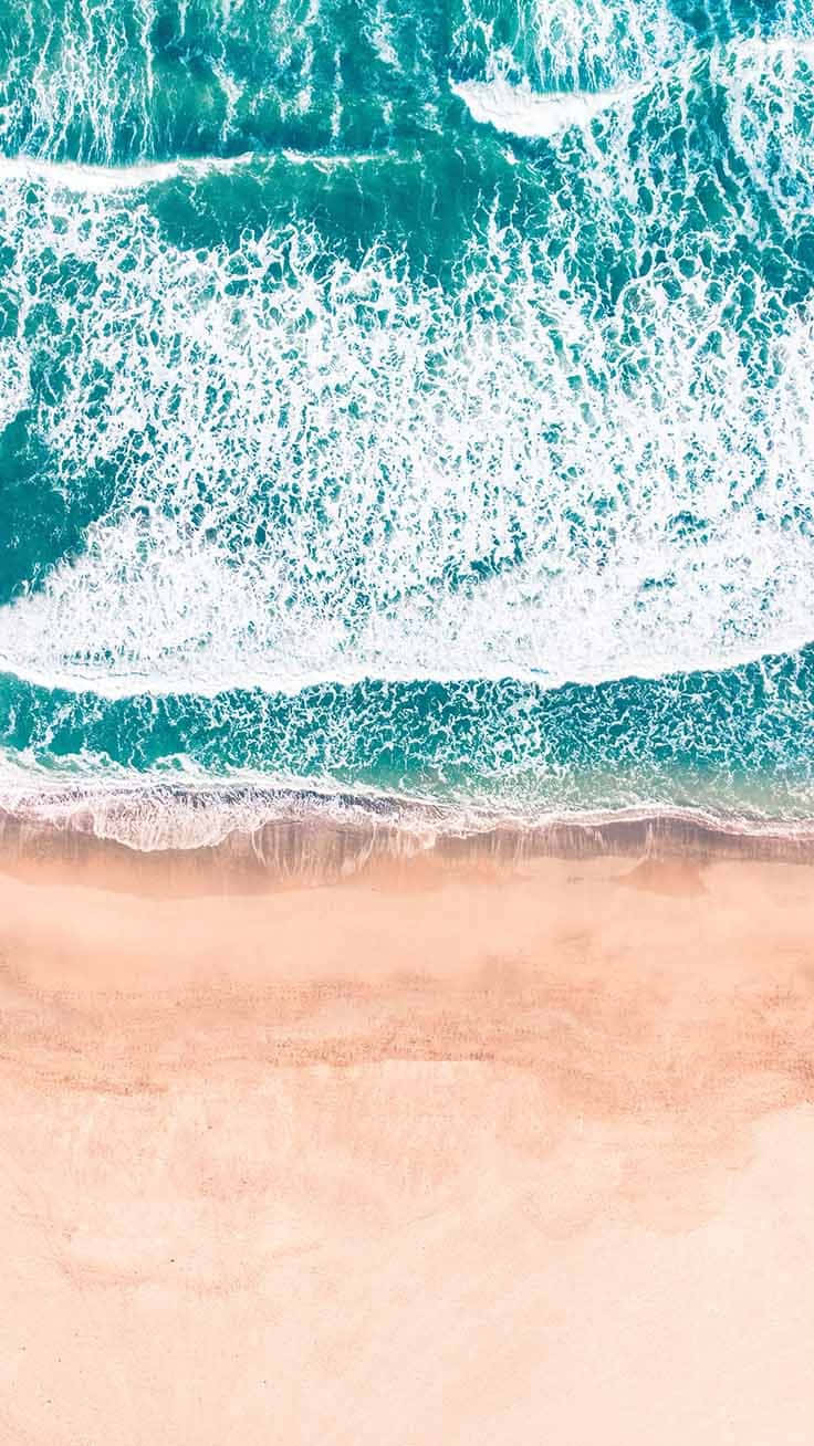 以時尚iPhone點亮你的夏日 壁紙 — 從高空俯瞰，綠松石色的海浪拍打著柔軟的蜜桃色沙灘。