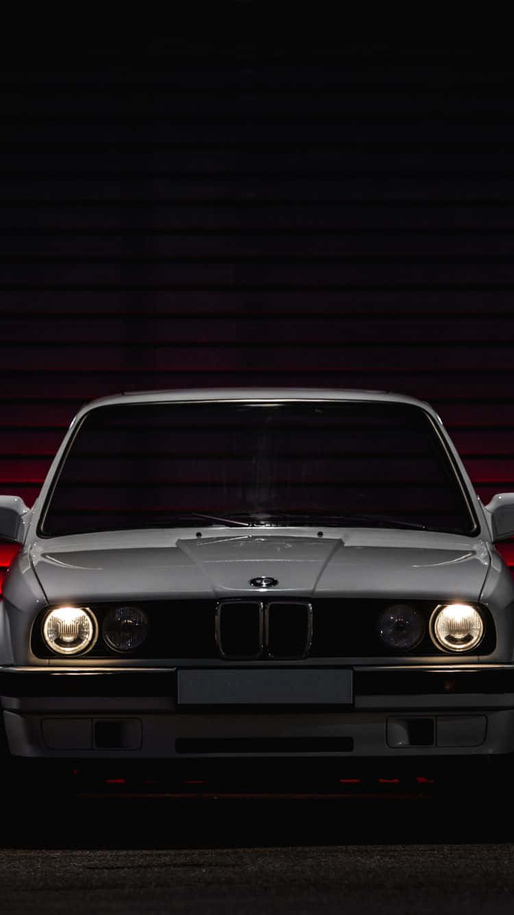 下載經典汽車iPhone壁紙，重溫懷舊風情！ 壁紙 — 一輛銀色BMW E30跑車，在昏暗光線下，亮著車頭燈，面向深紅色條板牆。