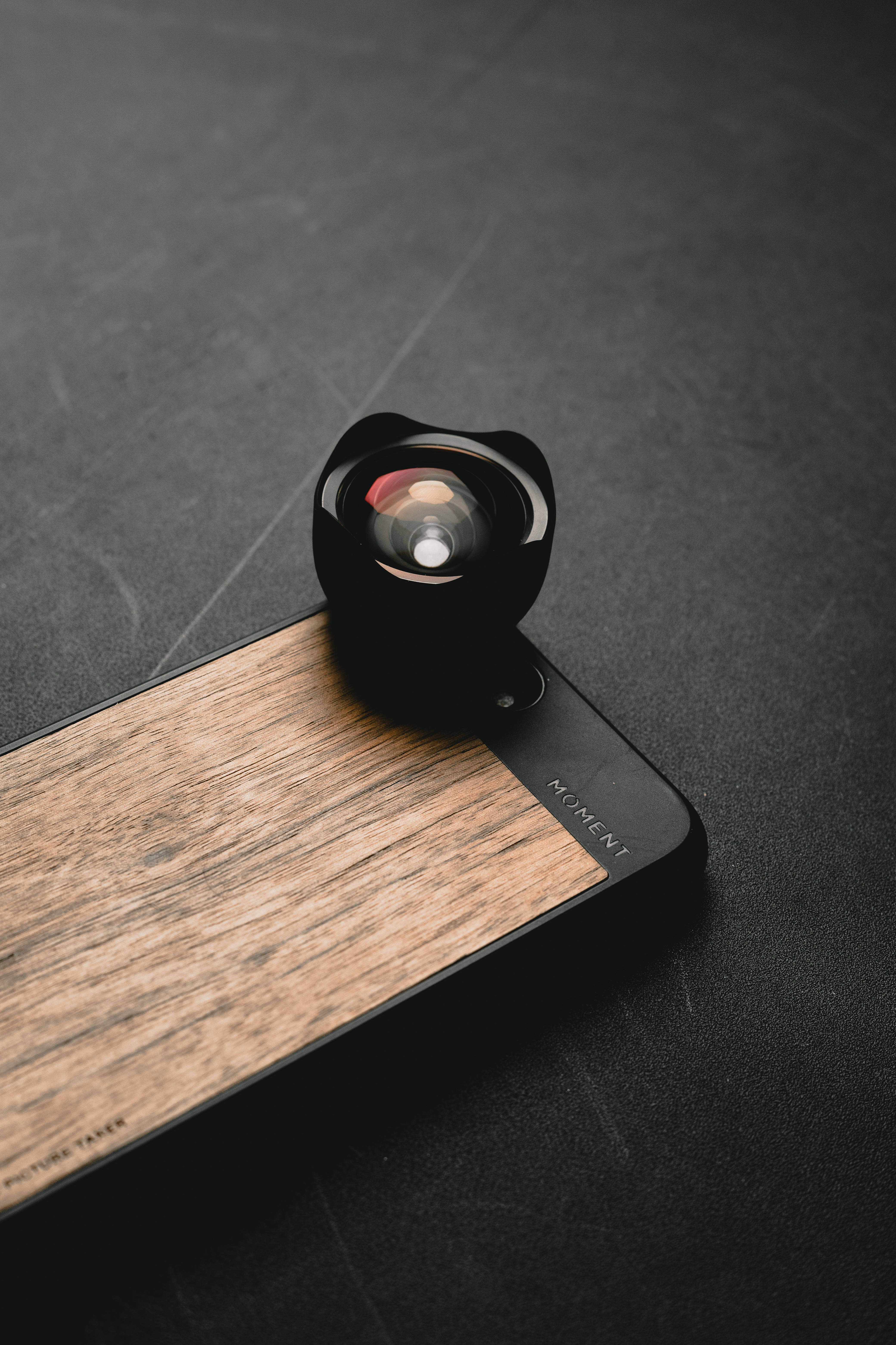 iPhone廣角鏡頭 壁紙 — 一個黑色魚眼鏡頭放置在木背手機殼上，置於深色紋理表面。