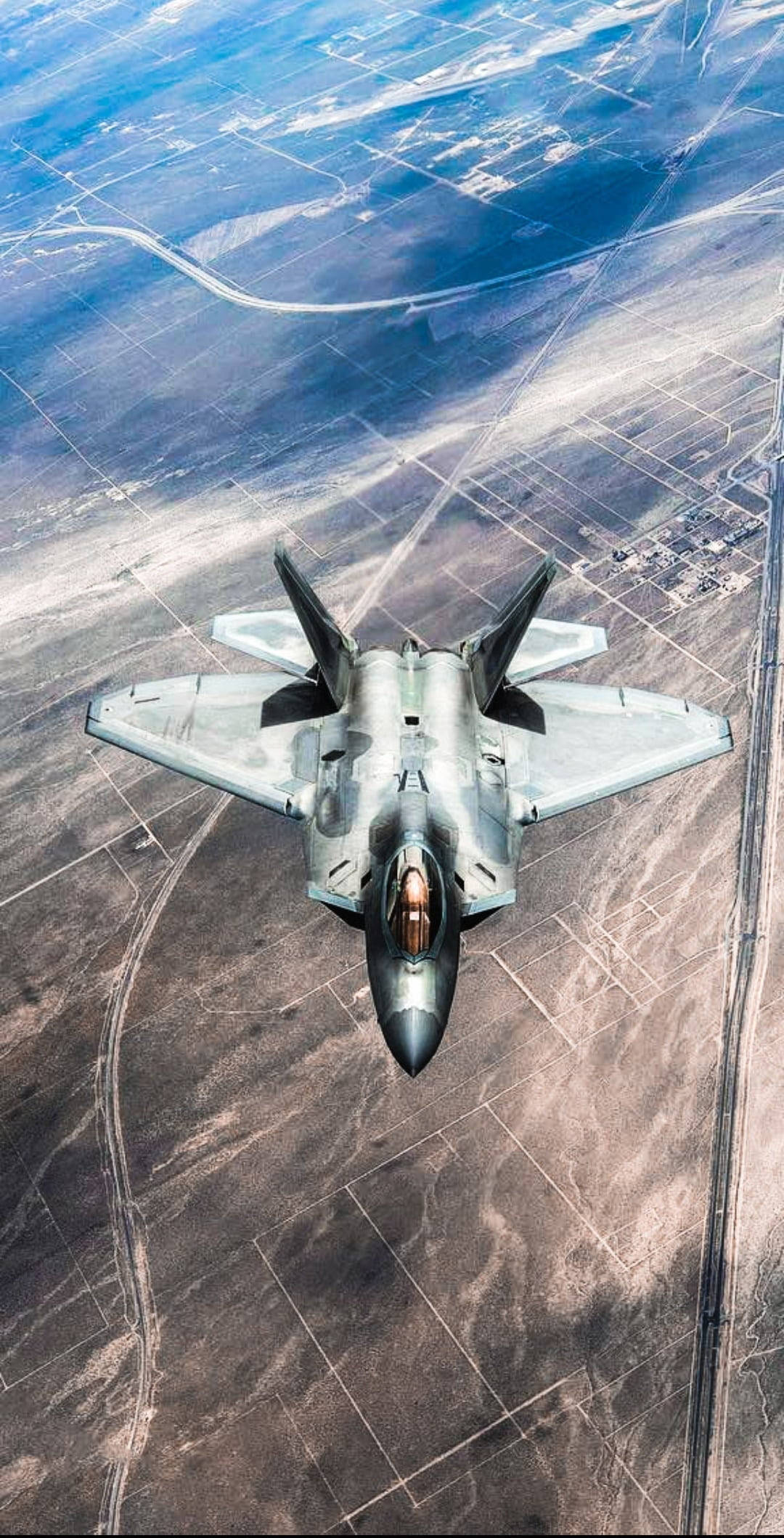 飛行灰色戰鬥機 iPhone 壁紙 — 一架流線型戰鬥機在幾何形狀的田野和蜿蜒道路的沙漠景觀上空翱翔。