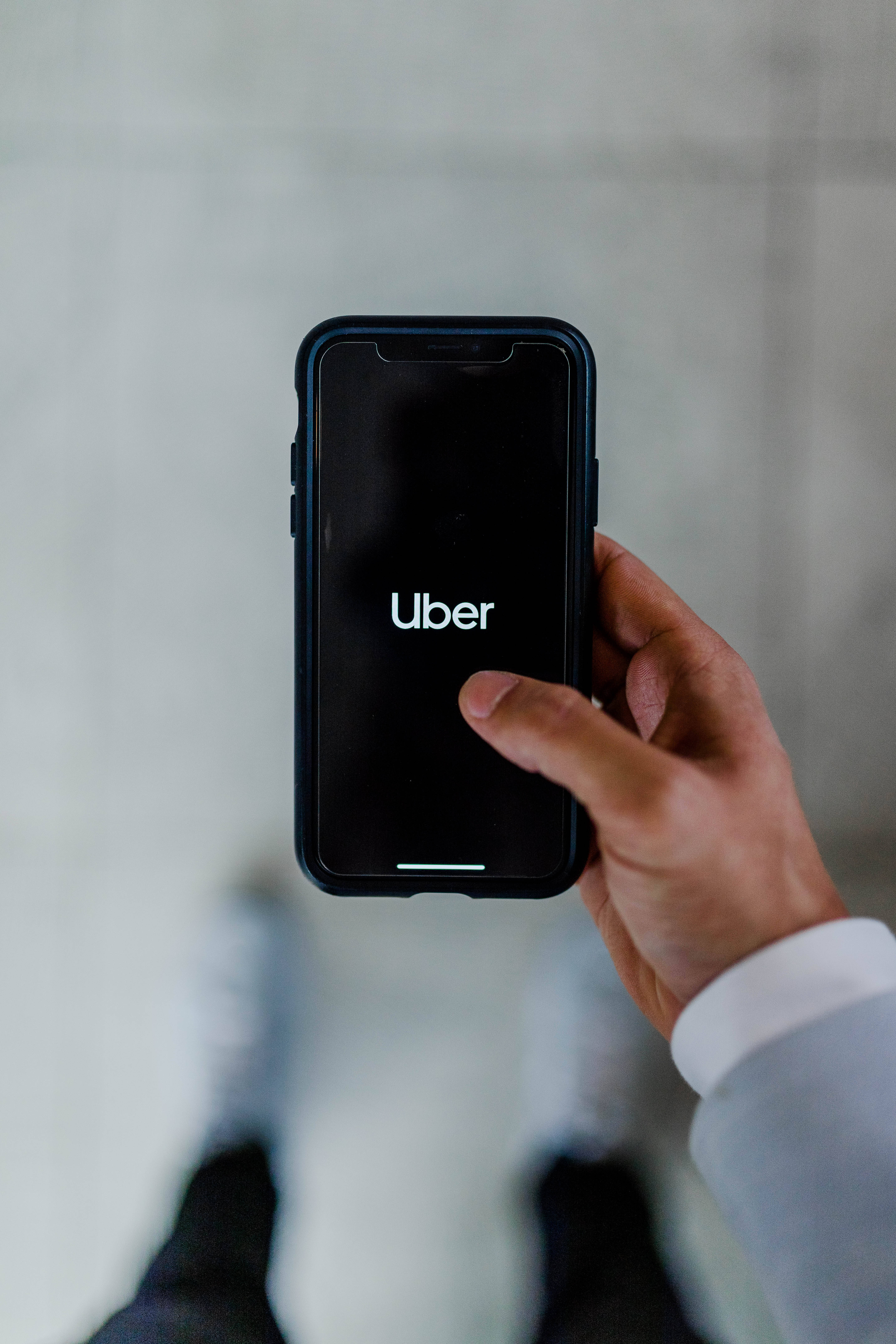 黑色iPhone iOS 13 Uber應用程式 壁紙 — 一隻手拿著智能手機，屏幕上顯示Uber應用程式標誌，背景為模糊的混凝土。