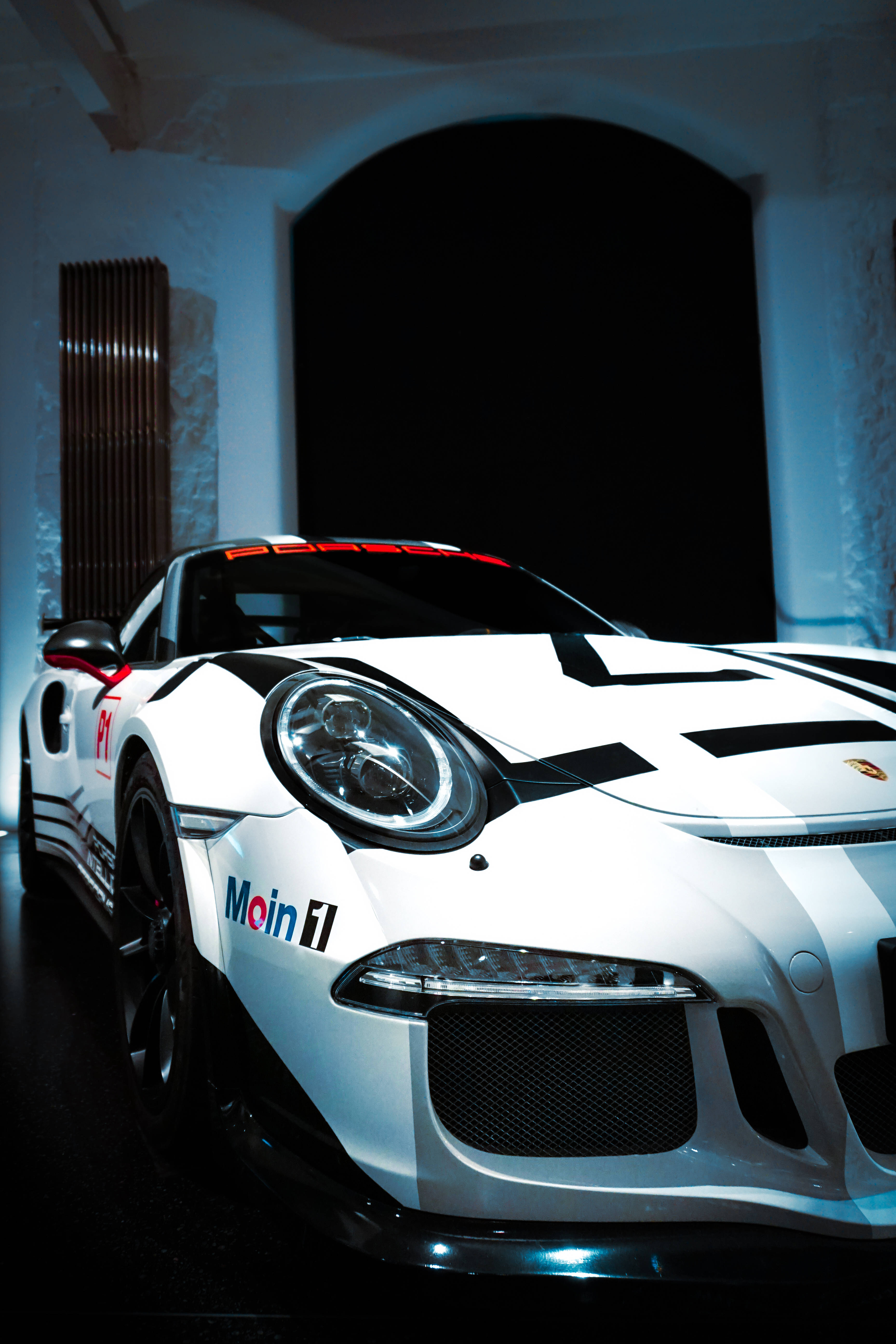 暗黑風格 iPhone 汽車 壁紙 — 一輛時尚的白色保時捷賽車，帶有黑色條紋和'Main F1'品牌標誌，在光線昏暗、拱形建築的車庫中戲劇性地被照亮。