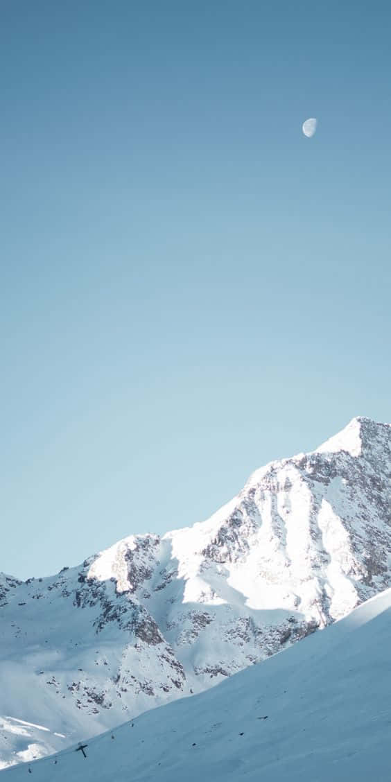 雪落白 iPhone 美景 壁紙 — 在晴朗的藍天下，一座積雪的山峰寧靜地聳立，可見一彎新月。